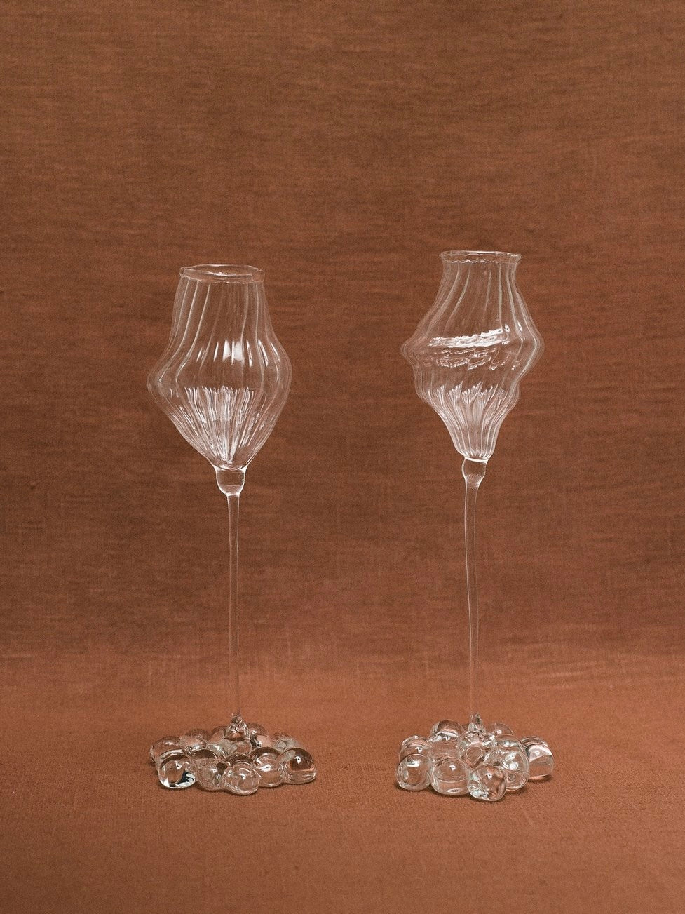 On a brown surface, two exquisite Orphée Liqueur Glasses from Justine Menard are showcased. These tall, mouth-blown glasses feature unique artistic forms with a textured and wavy design. Each handmade piece rests on a base embellished with multiple glass pebbles. The background is draped in a matching brown fabric that enhances the overall aesthetic.