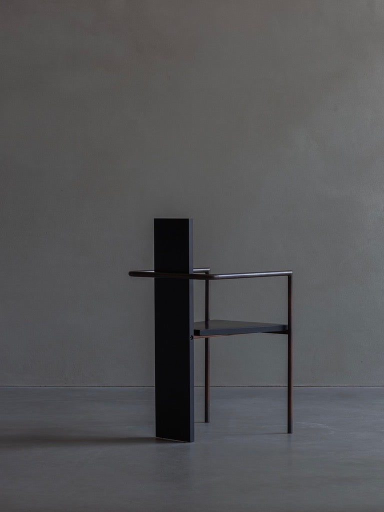 The Jonas Bohlin - CONCRETE - black by Bicci de' Medici stands elegantly against a gray backdrop, showcasing its minimalist design with sleek angular lines and a single armrest. Its artistic silhouette merges seamlessly with the smooth concrete floor.