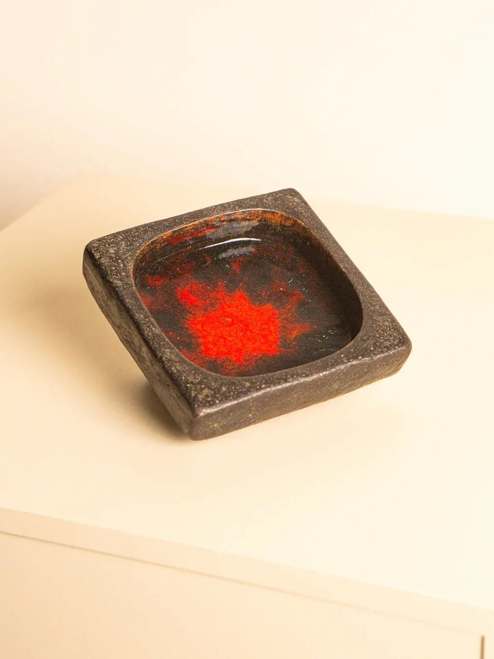 A 60's Square Ceramic Tray by Jan Van Erp, featuring a square design with a dark stone finish and a glossy red interior, reminiscent of a decorative work of art, rests at an angle on a light-colored surface under warm lighting. This elegant piece is part of the Treaptyque brand collection.