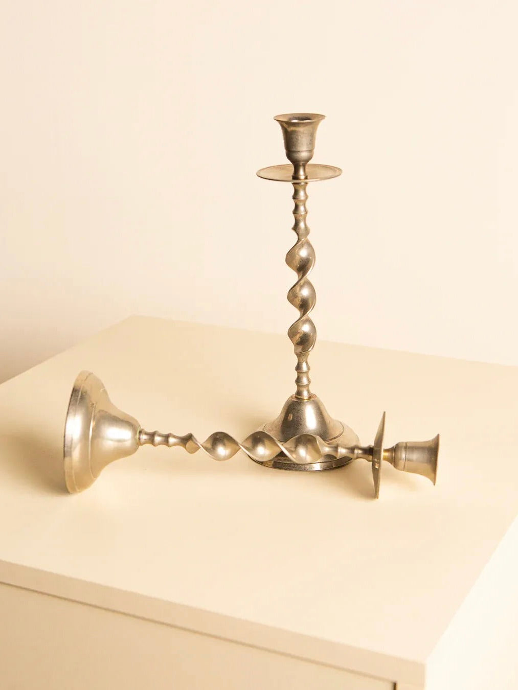 A pair of 60's Italian Metal Candleholders from Treaptyque, featuring twisted bodies and spiral designs, are displayed on a light-colored surface. One candleholder stands upright while the other rests on its side against a plain, off-white wall that evokes classic 60s decor.