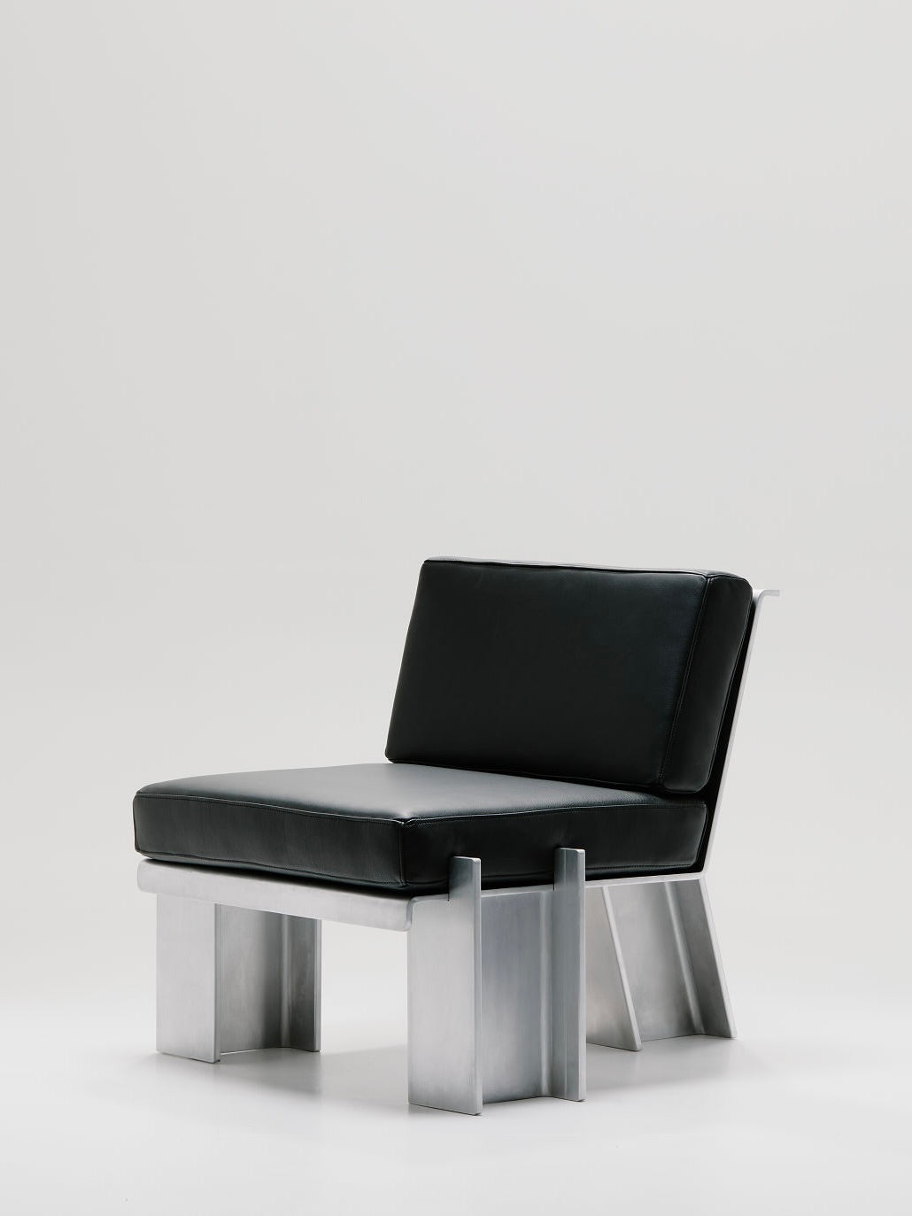 The Beam Lounge Chair by Marquel Williams boasts a sleek design with a black cushioned seat and full-grain leather backrest. Its distinctive metal frame and waxed aluminum legs create a contemporary, industrial aesthetic against a plain white background.