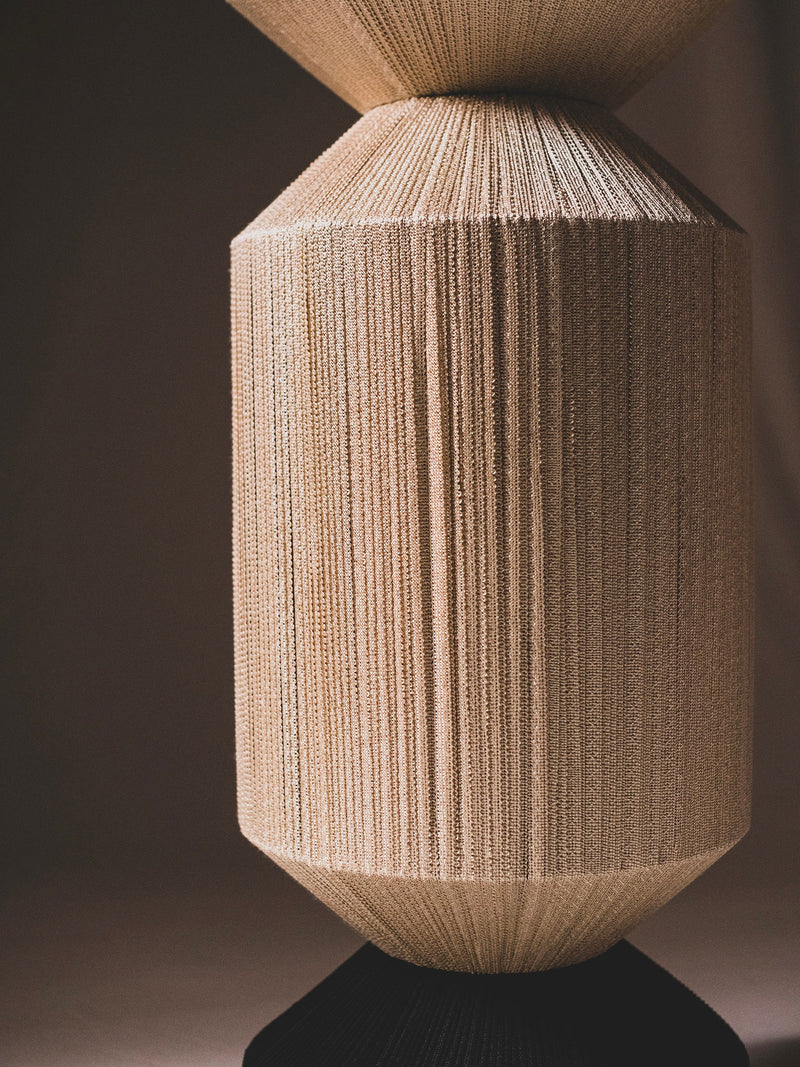 The Column IX Sculpture by ELIURPI is a cylindrical wooden artwork with intricate handmade trimmings, vertical grooves, and a textured surface. The lighting enhances the wood's natural tones against a dark backdrop.