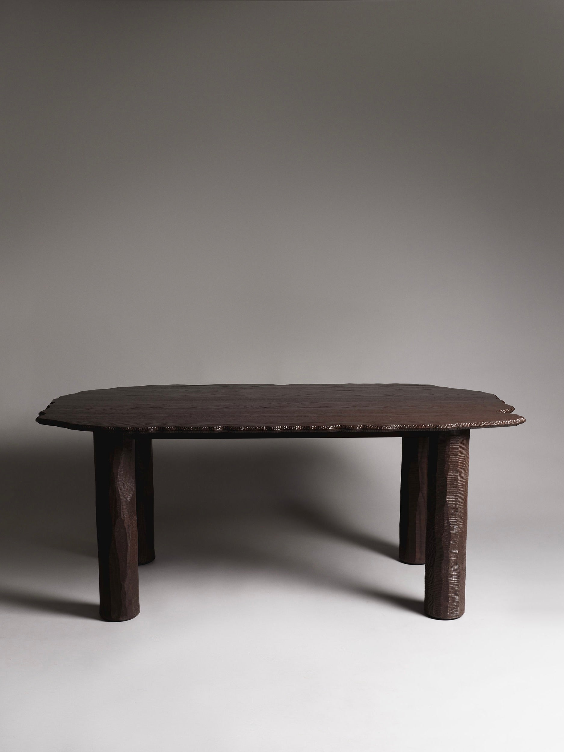 The Mare Dining Table by Ombia Studio features a dark wood, slightly oval top and sturdy cylindrical legs. It's hand-carved from exquisite wood and set against a plain gray background, casting subtle shadows beneath.