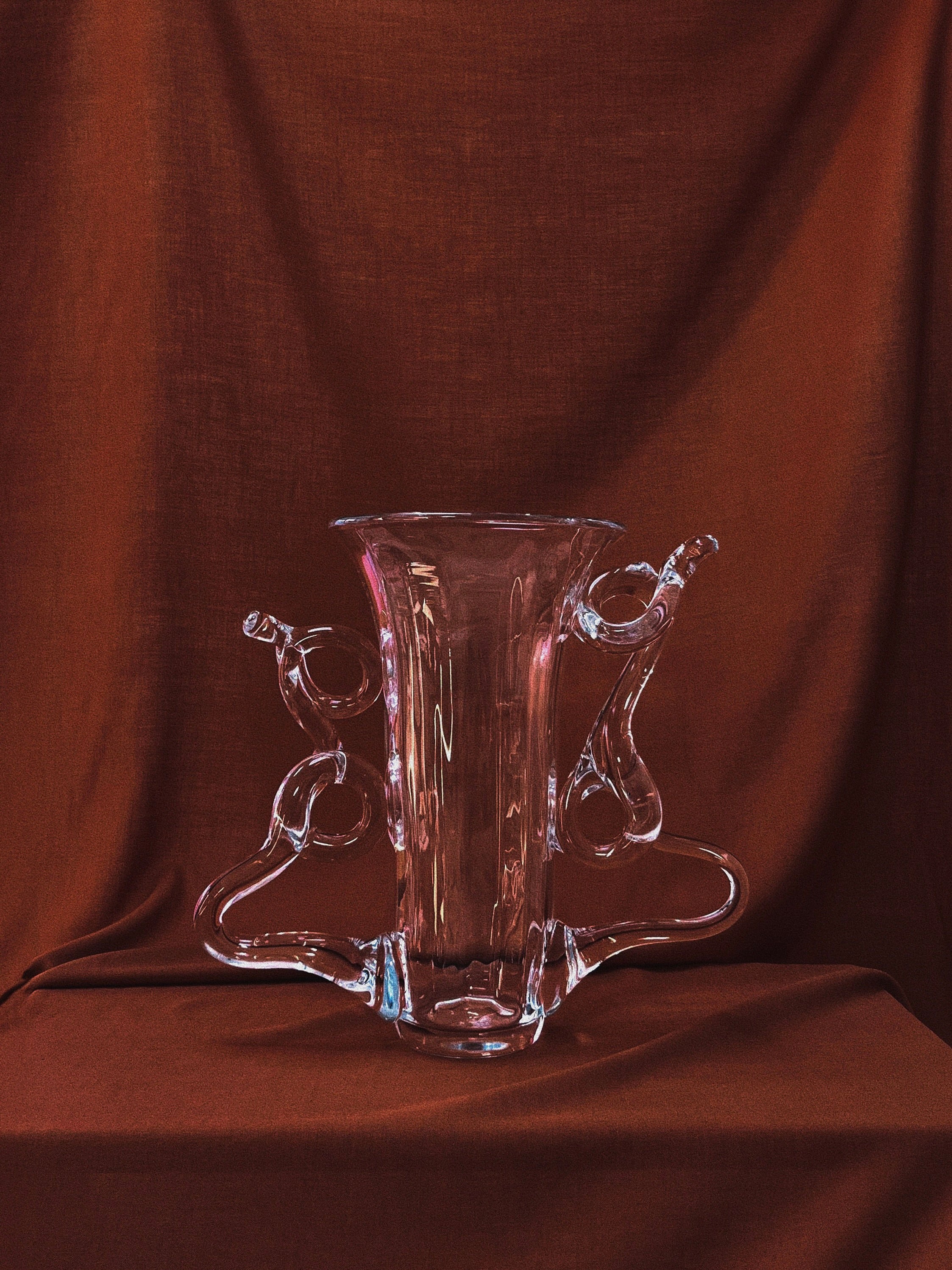 The "Vine" Vase by Hors-d'Oeuvre, a clear glass creation with intricately swirling handles, is showcased against a rich brown fabric backdrop. Handcrafted with elegance, it casts subtle reflections and shadows that emphasize its artistic curves and exceptional craftsmanship in a sculptural context.