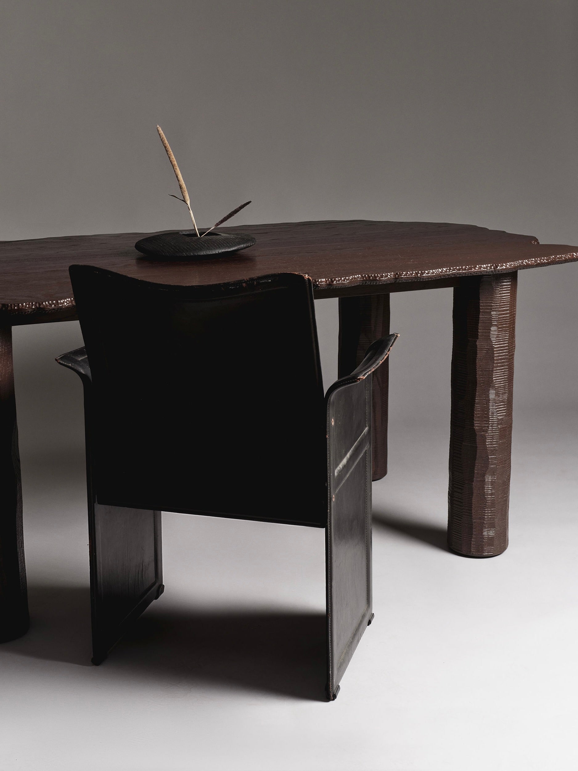 The Mare Dining Table by Ombia Studio is a rustic, handcrafted wooden table with an irregular shape and textured legs, paired with a dark wooden chair. Its round base features a carved feather detail, adding artistic flair to any minimalist setting.