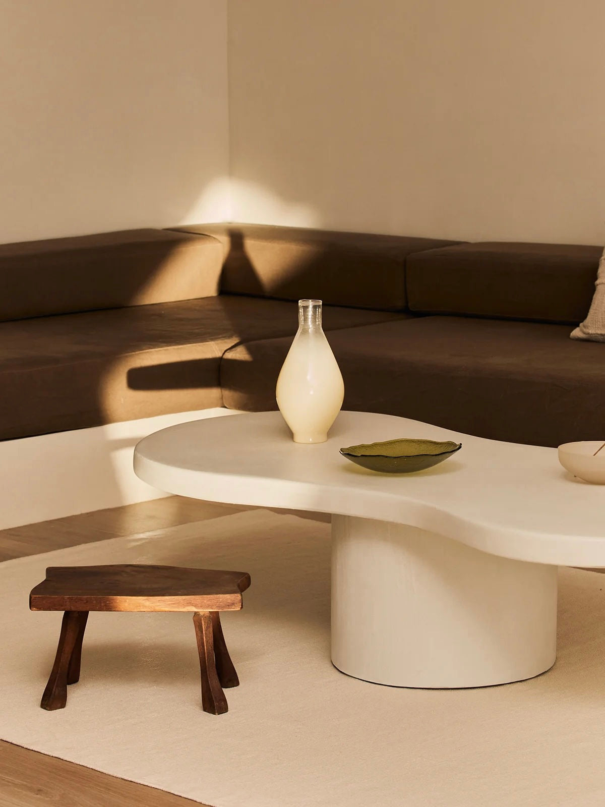 A minimalist living room features the "Pesto" coffee table by MarlotBaus, elegantly showcasing a vase and a small dish. A brown low sofa lines the background, while an artisan-crafted wooden stool rests on a cream rug. Soft, warm lighting creates gentle shadows throughout the inviting space.