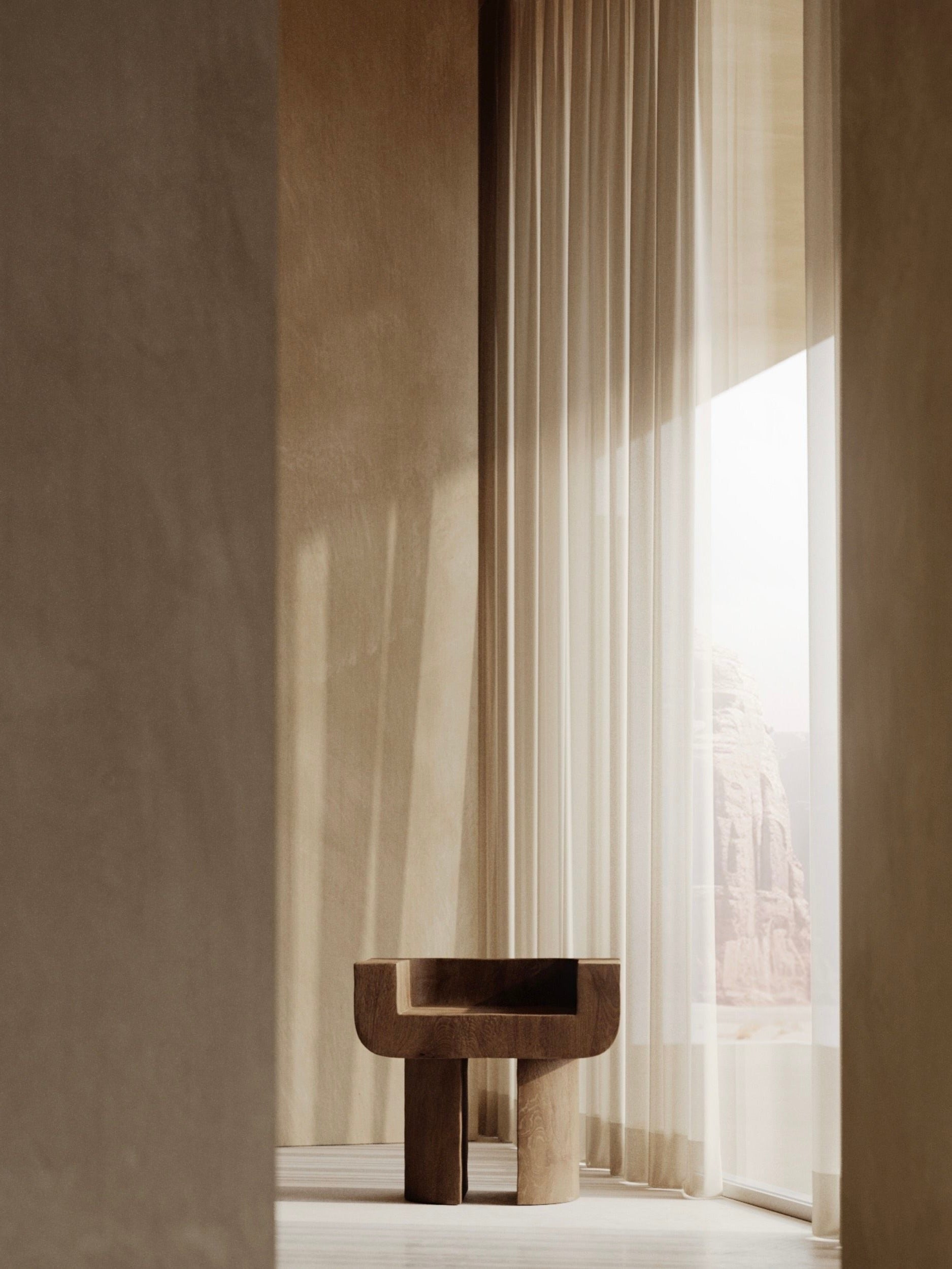 A minimalist interior features a Monolith M_001 Chair with a throne-like form by a floor-to-ceiling window. Sheer curtains diffuse soft light, illuminating neutral-toned walls and enhancing the serene ambiance.
