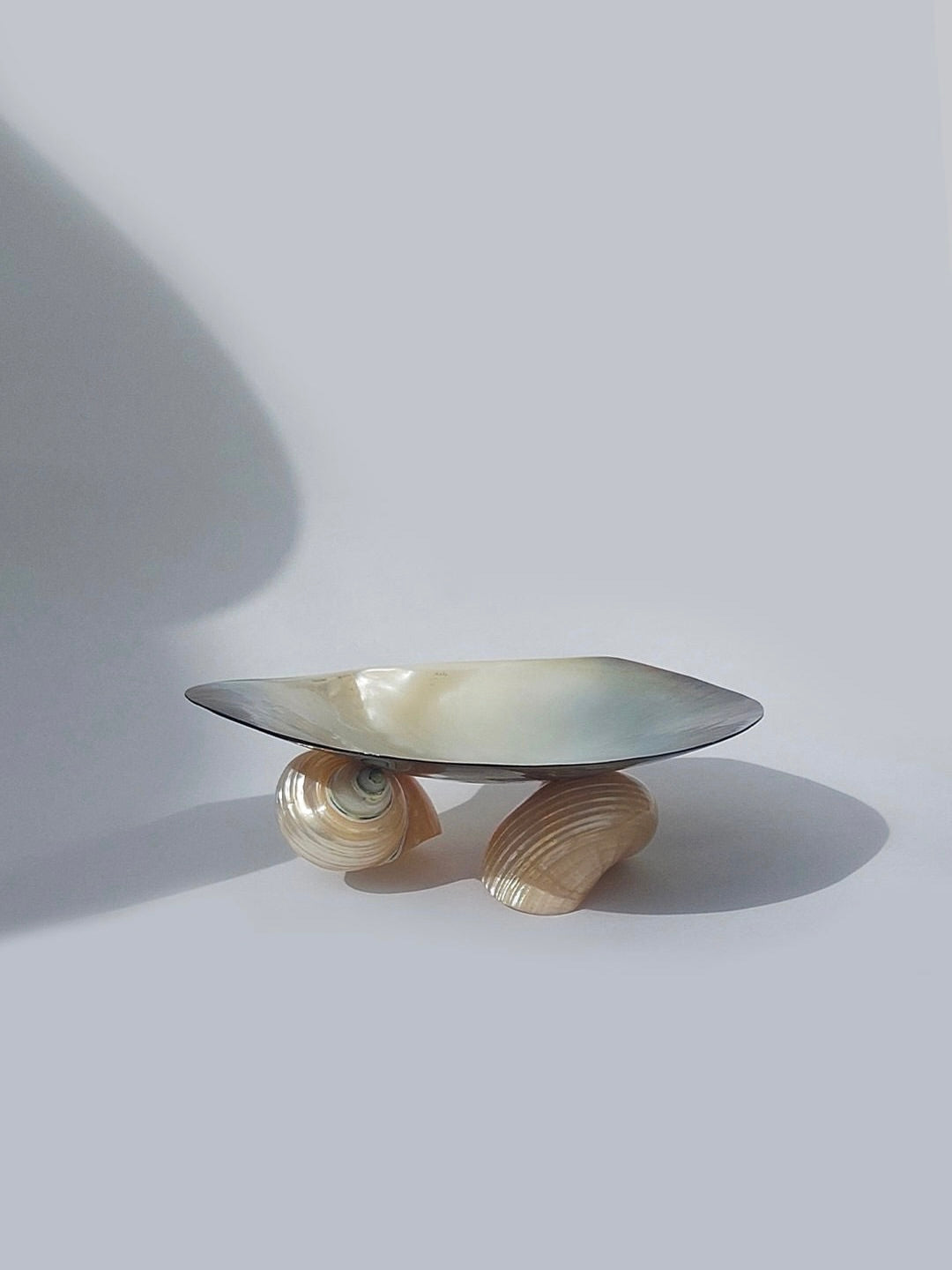 A modern, artistic display titled the Mother-Of-Pearl Vide Poche by Les Objoies rests atop three elegantly curved supports reminiscent of shells. This decorative piece showcases a sleek, metallic finish complemented by a background gradient of light and dark grey, casting subtle shadows that highlight its unique design and mother-of-pearl undertones.