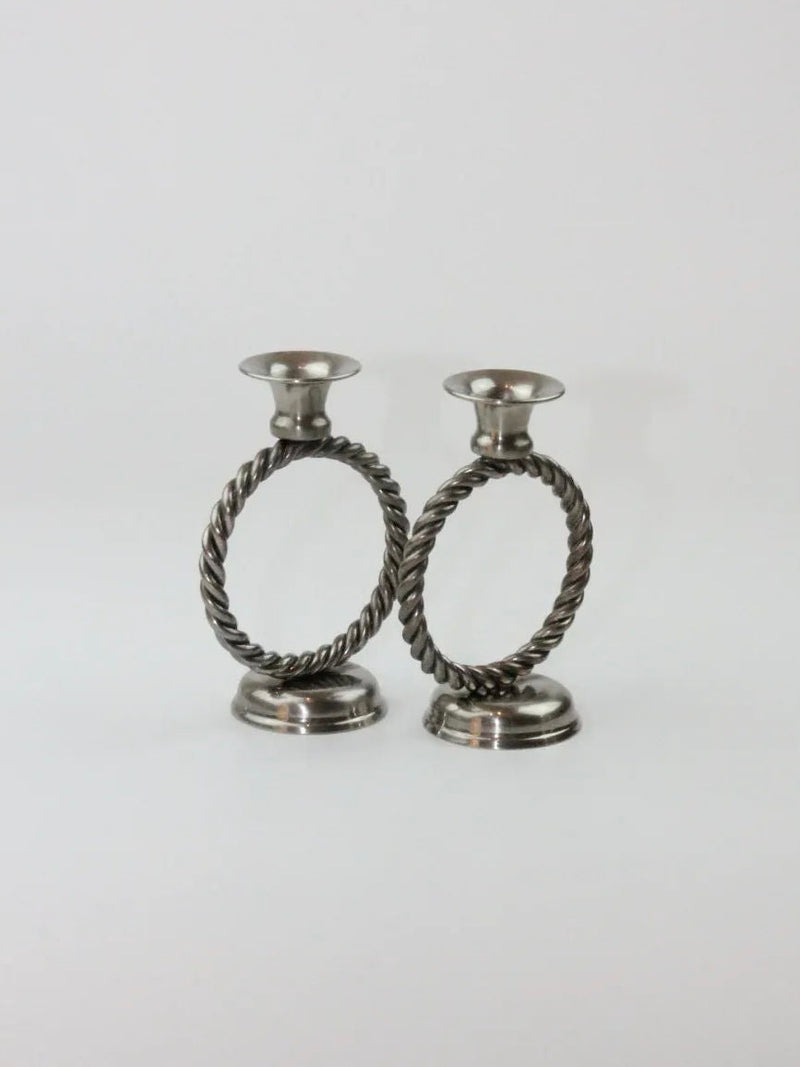 Introducing the Mental Candle Holders from Boga Avante Shop, a pair of classic and sophisticated silver candle holders featuring elegant twisted circular designs. Each metal holder is designed with a round base and a cup at the top for placing candles. Set against a plain, light-colored background, their timeless design truly stands out.