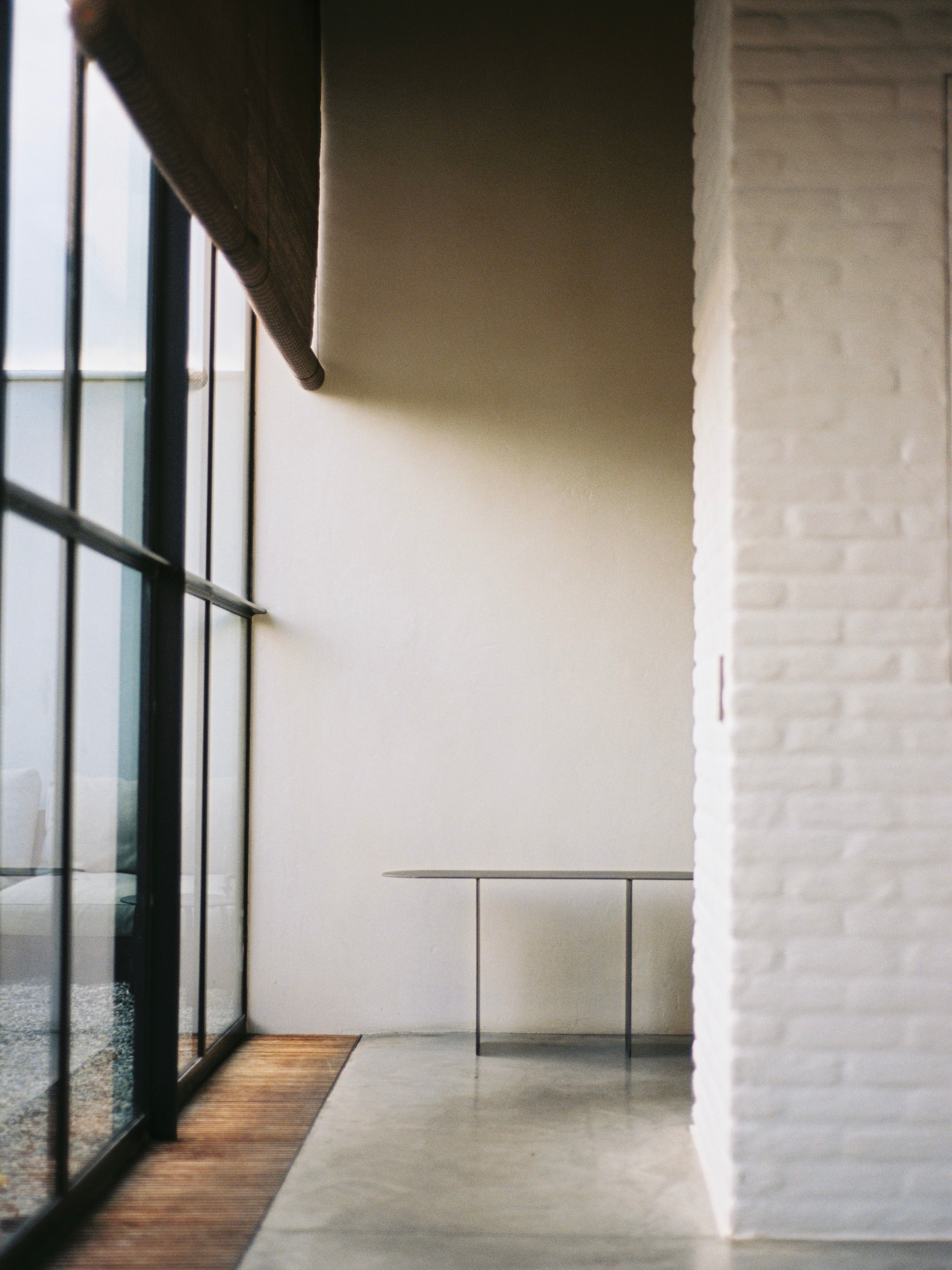 The minimalist interior features a white brick wall, large floor-to-ceiling windows, and a small table. "The Altar," a limited edition prayer kneeler by COSEINCORSO, adds an elegant touch, while natural light streams in through the windows, creating a serene and airy atmosphere.