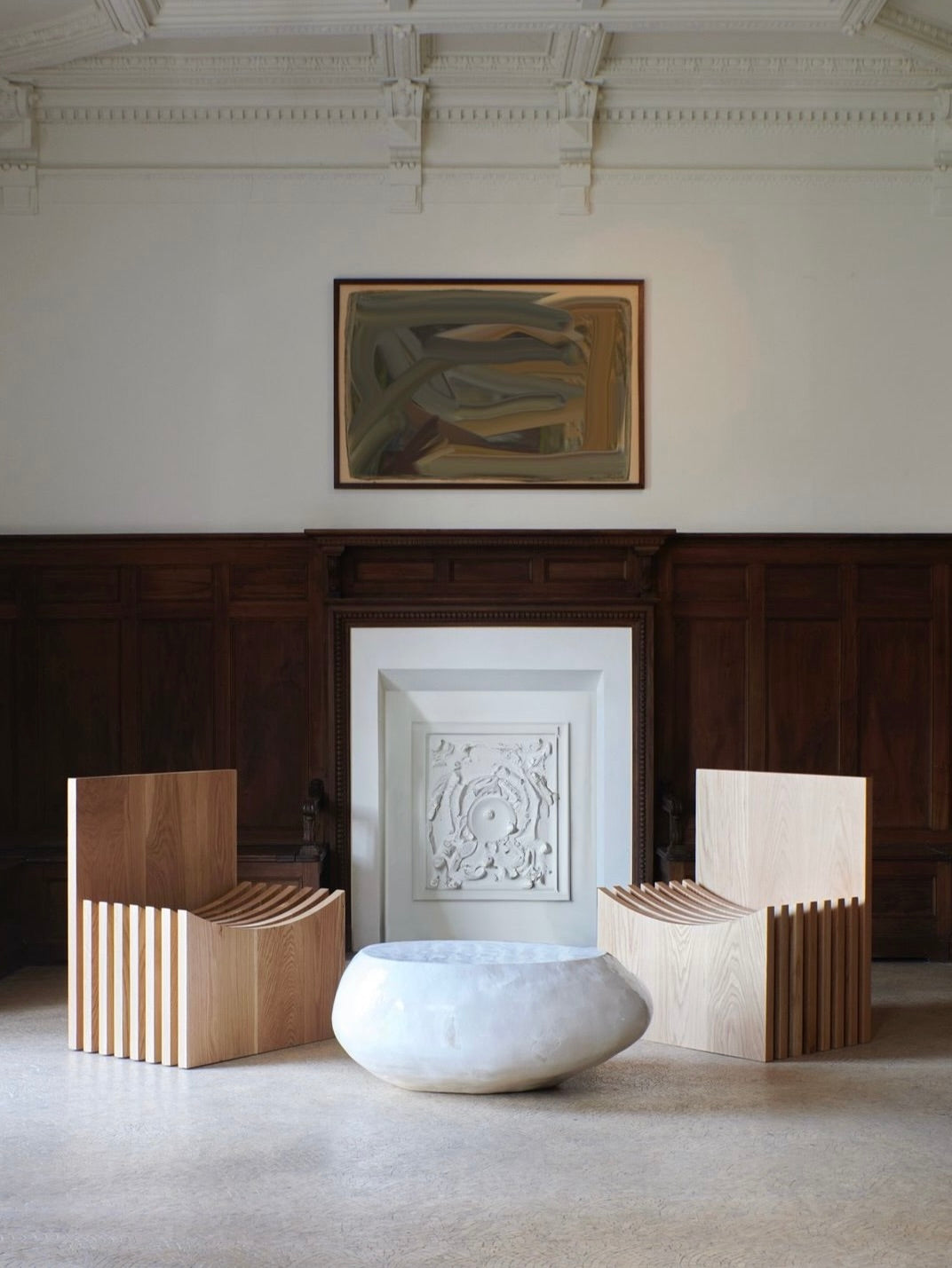 A modern living room showcases a minimalist design with two hand-crafted Melides Chairs from Project 213A, made from solid oak, positioned on either side of a large, round marble coffee table. The room is adorned with intricate wall moldings, a decorative fireplace, and features a framed abstract painting above.