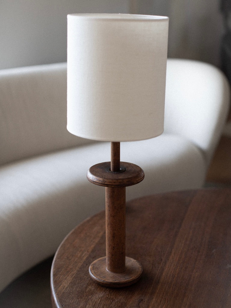 A Midcentury Table Lamp by Out For Lunch, featuring a cylindrical white lampshade and a wooden handle, is placed on a circular wooden table. In the background, a curved white sofa enhances the mid-century modern interior design.