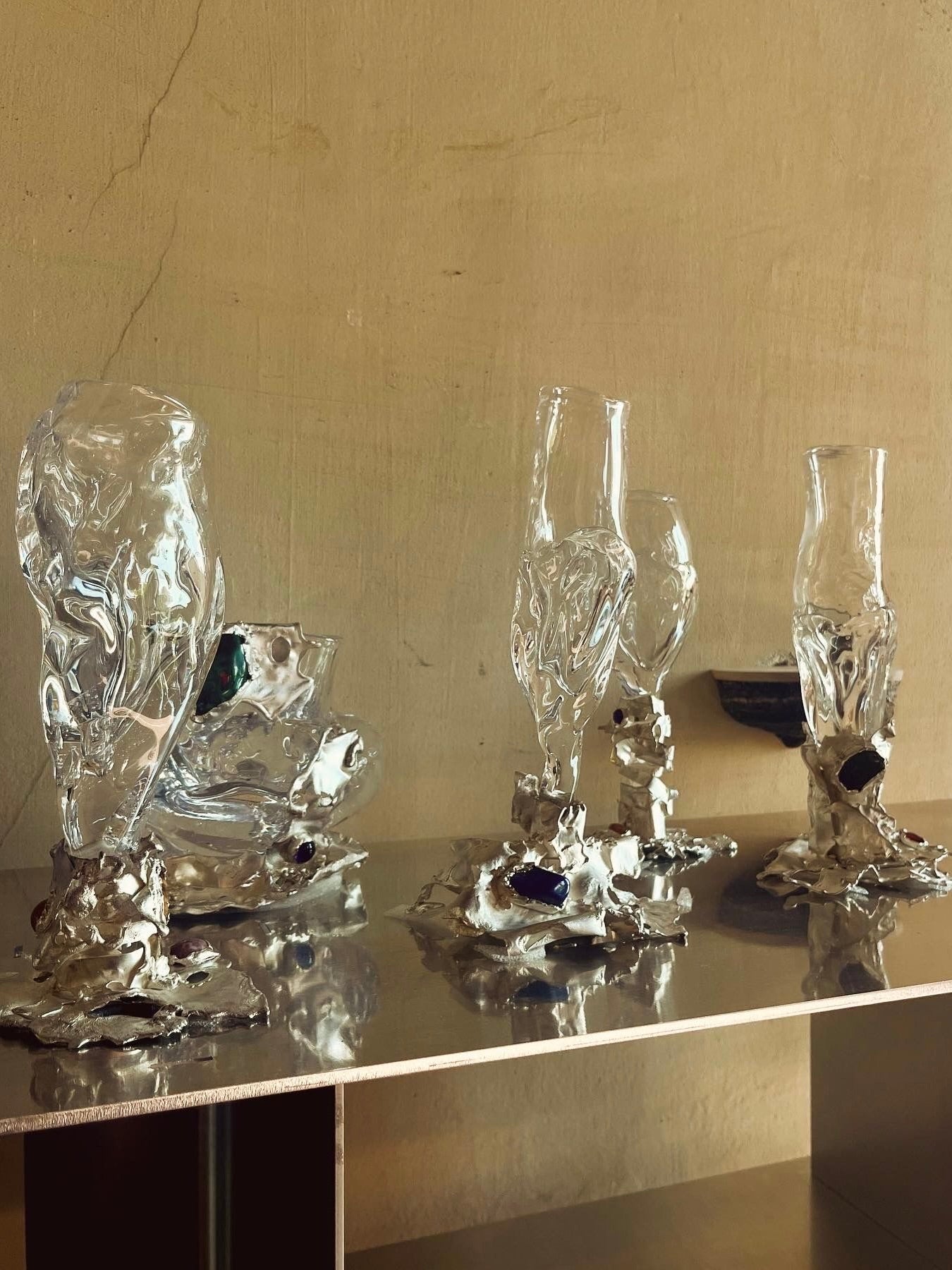 A display of five Jewelled Goblet no. 5 artworks by Lindsey Fontijn from Szkło Studio features abstract, hand-blown glass pieces with fluid, distorted forms arranged on a reflective surface against a neutral beige wall. Each piece is mounted onto a rough silver-colored base, reminiscent of goblets with silver stems, enhancing the avant-garde aesthetic.