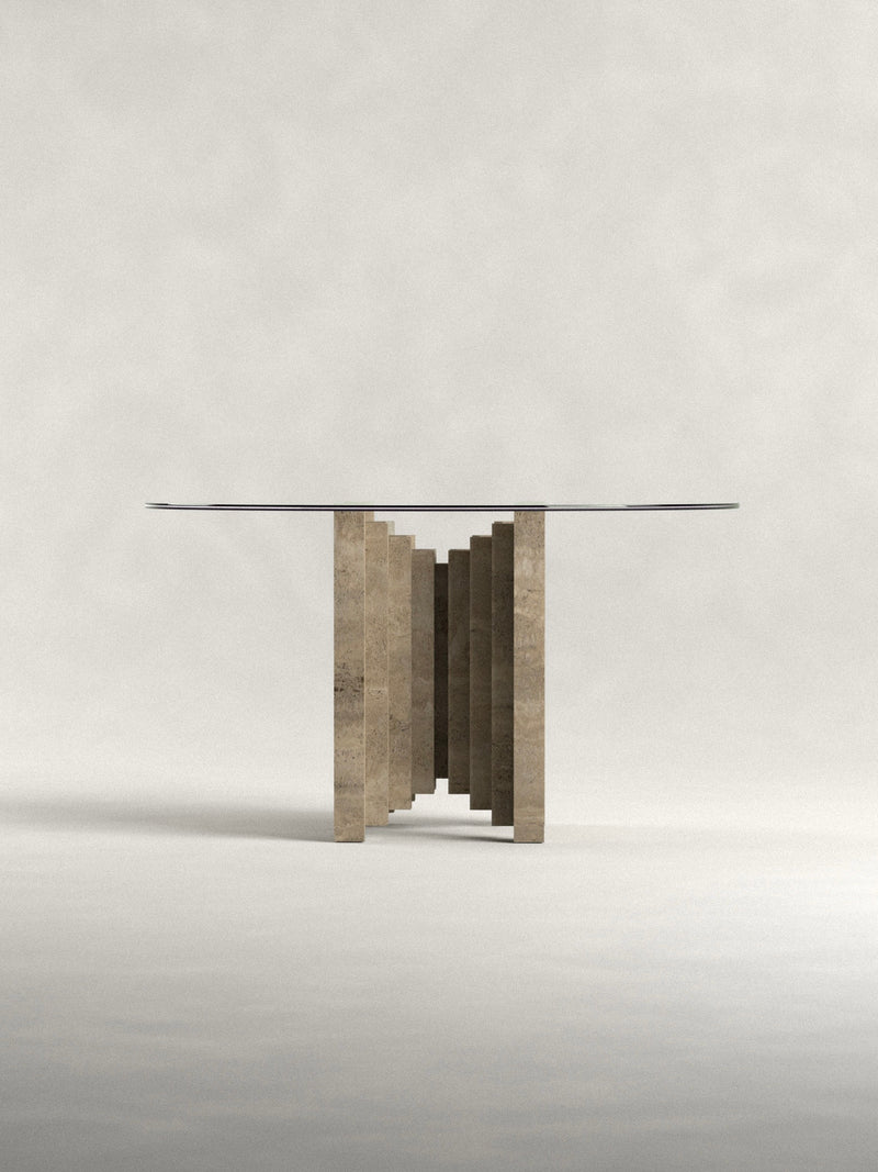 The 'Calakmul' Dining Table S2 by Siete Studio features a sleek glass top and a handcrafted geometric travertine base with rectangular slabs in a zigzag pattern, offering elegance and minimalist design against a neutral background.