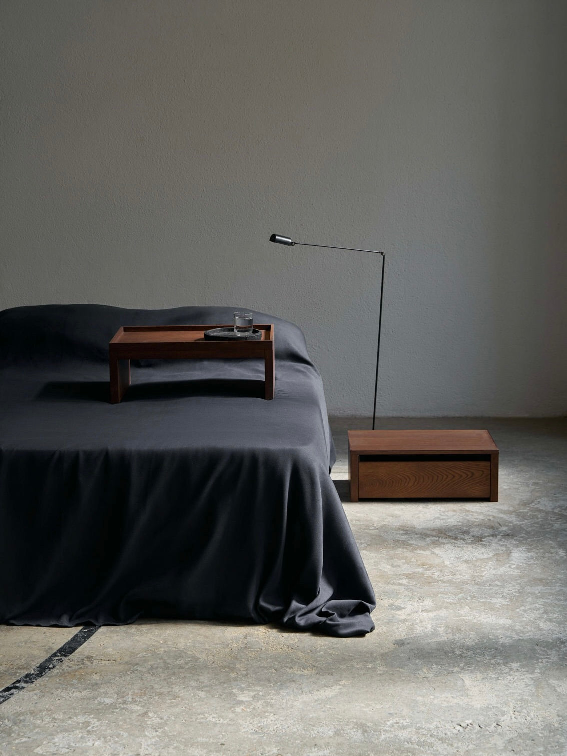 A minimalist bedroom showcases a dark, low bed with a flowing black duvet. The ASERIES OF OBSTACLES, SL Eileen Bedside Table + Tray features a glass and small rectangular lamp. A concrete floor completes the room's modern neutral palette.