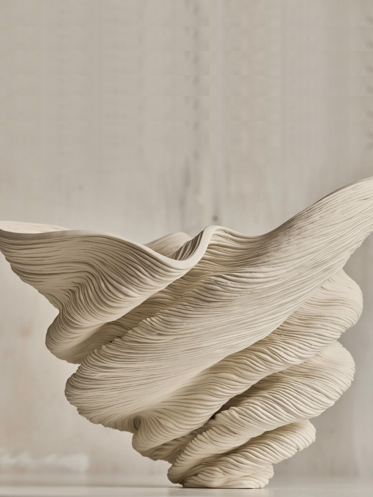 The 'Medium Flutter' by The Soucy Shop is a unique beige ceramic sculpture handcrafted with porcelain coils, featuring an intricately textured design of swirling, layered waves and a dynamic twisted form set against a neutral background.