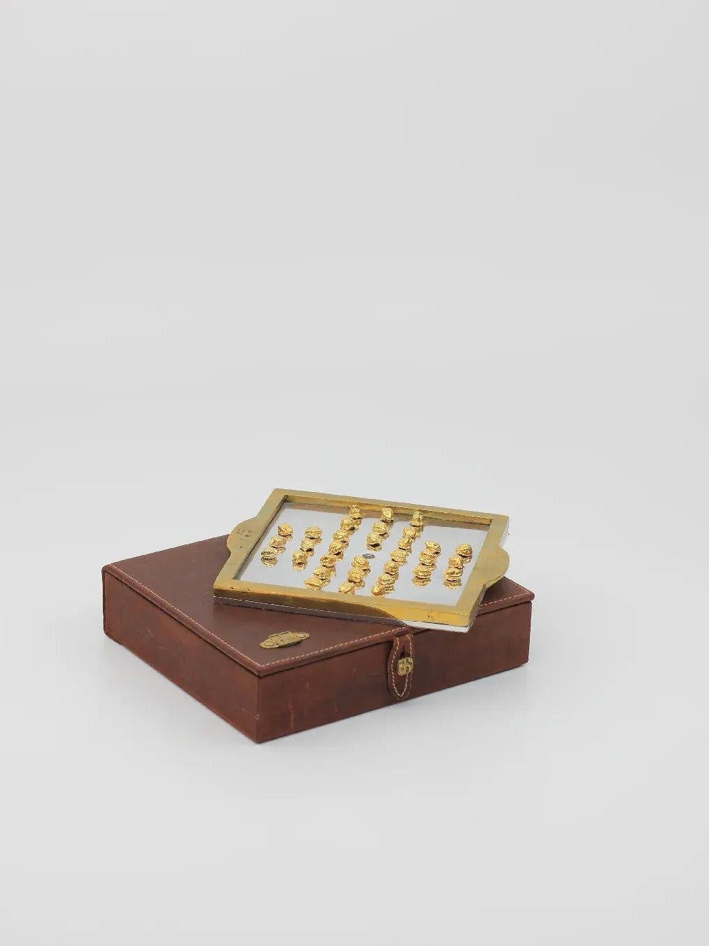 An exquisite tray of gold figurines is elegantly displayed on a closed brown leather box with a latch, reminiscent of vintage designs, all set against a plain white background, paying homage to the English Solitaire crafted by Boga Avante Shop.