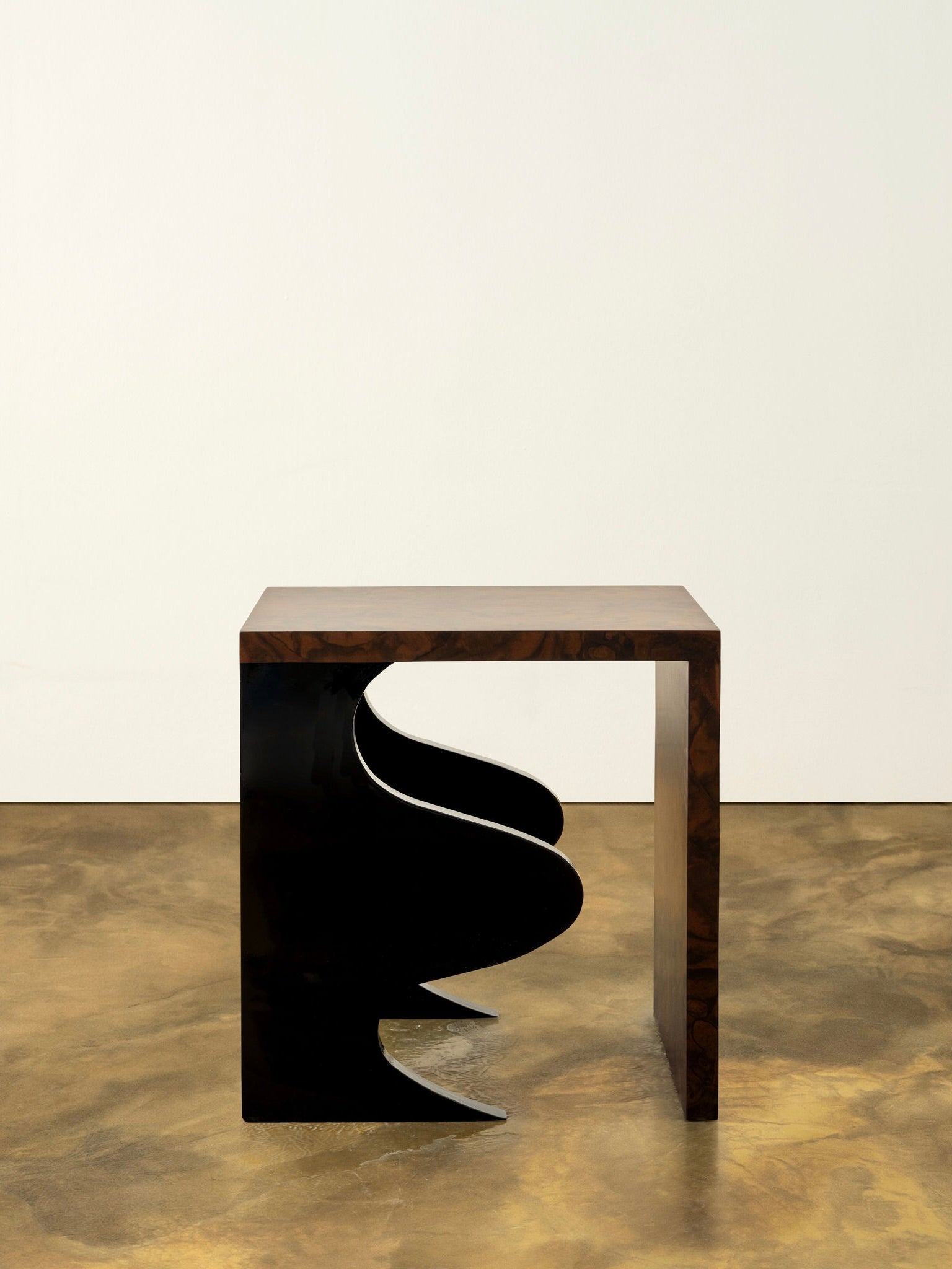 Introducing the Harlequin Side Table by Animate Objects, a limited edition luxury furniture piece that features a sculptural design with an abstract black cutout element. The walnut burl veneer over gloss lacquered wood adds to its sophistication as it gracefully stands on a polished wooden floor against a plain white wall, showcasing its elegant craftsmanship in every handcrafted detail.