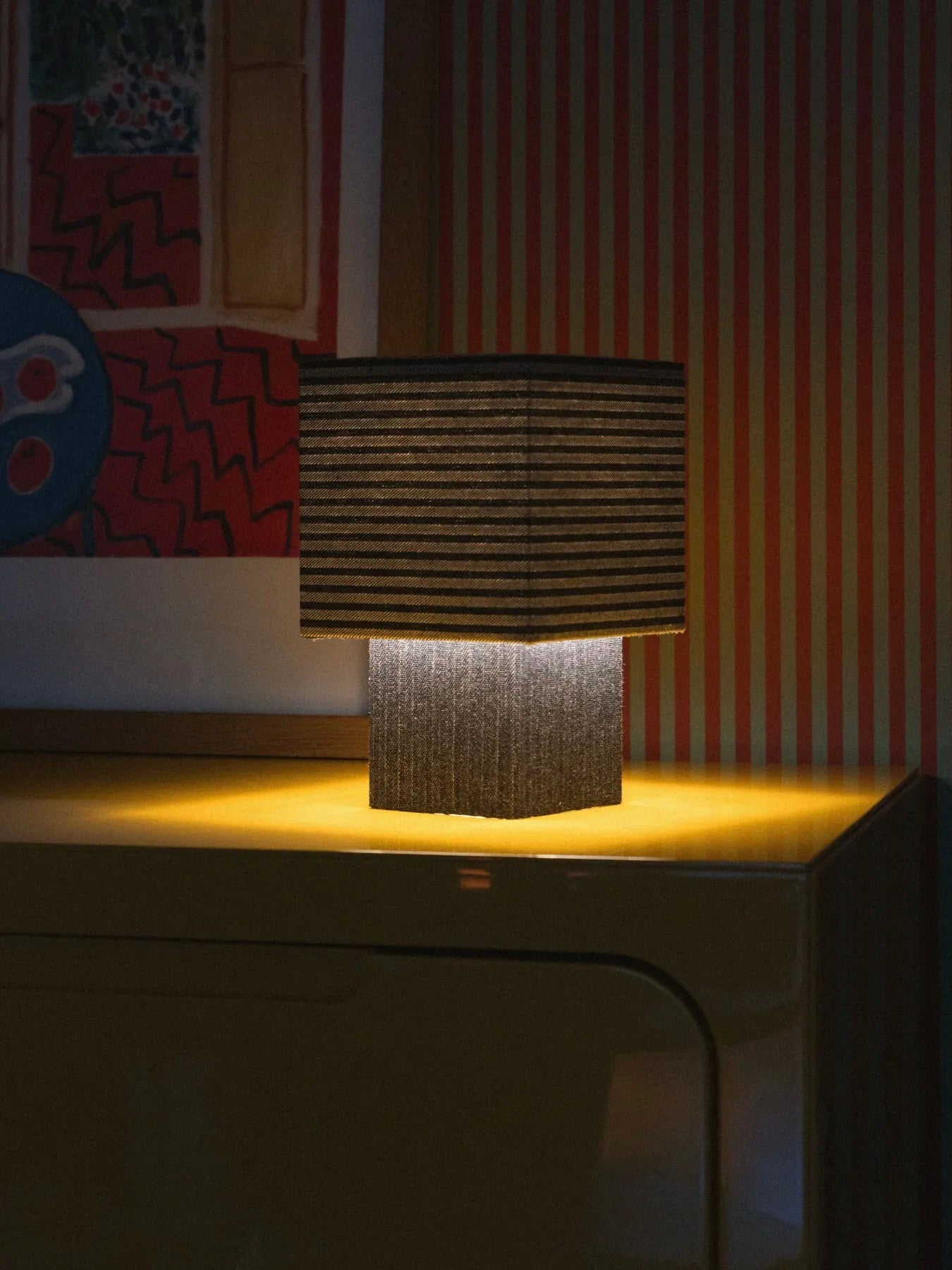 A softly glowing "La Petite Boîte" - Striped Denim Table Lamp by mattina moderna, featuring a square-shaped design and crafted from deadstock fabrics, rests on a yellow table. In the background, a framed abstract artwork bursting with various colors and patterns is set against a red and white striped wall.