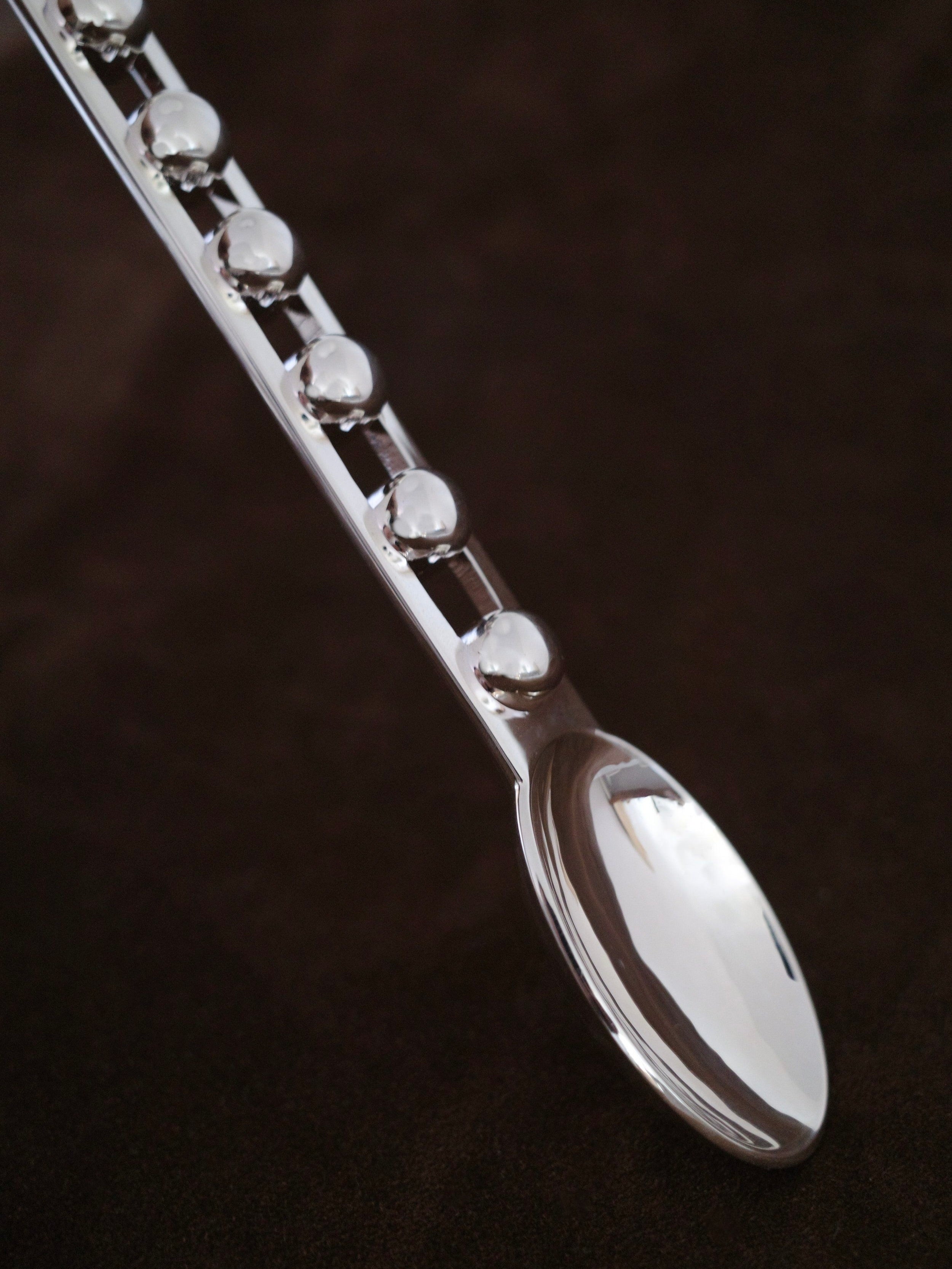 A close-up of a shiny, metallic spoon from the Jessi Burch "Seven Ball" Cutlery Set shows off its distinct handle design adorned with evenly spaced, raised spherical ornaments. The dark, solid background accentuates this piece of functional art.