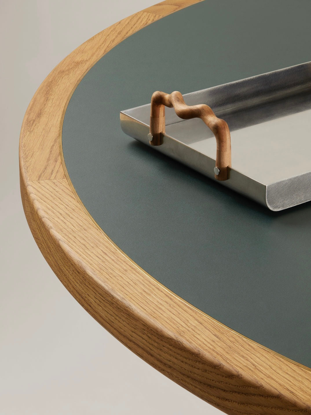 The Jack Table by HAUVETTE & MADANI showcases a circular design with a wooden border and a deep green tabletop, reminiscent of classic games tables. It includes an elegant rectangular metal tray adorned with wave-patterned wooden handles, perfectly merging minimalistic and contemporary aesthetics.