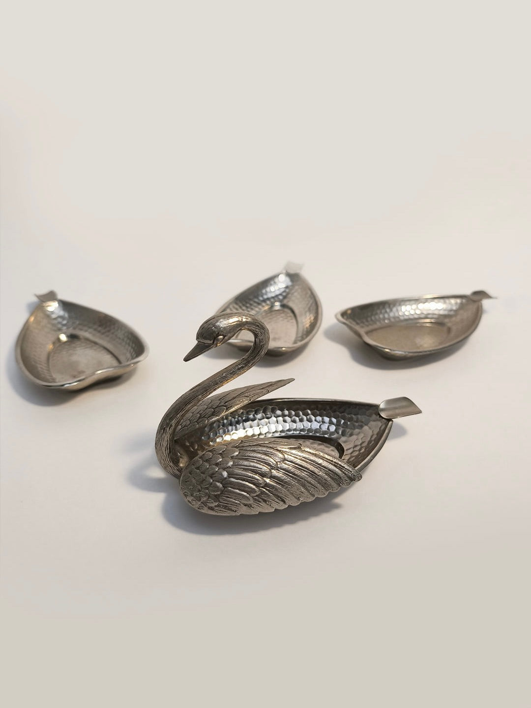 The Swan Ashtray by Les Objoies features an intricate silver swan dish that serves as a stunning metallic centerpiece ashtray. Accompanying this are three smaller swan-shaped dishes with detailed feather textures, each functioning as detachable ashtrays against a minimalist backdrop.