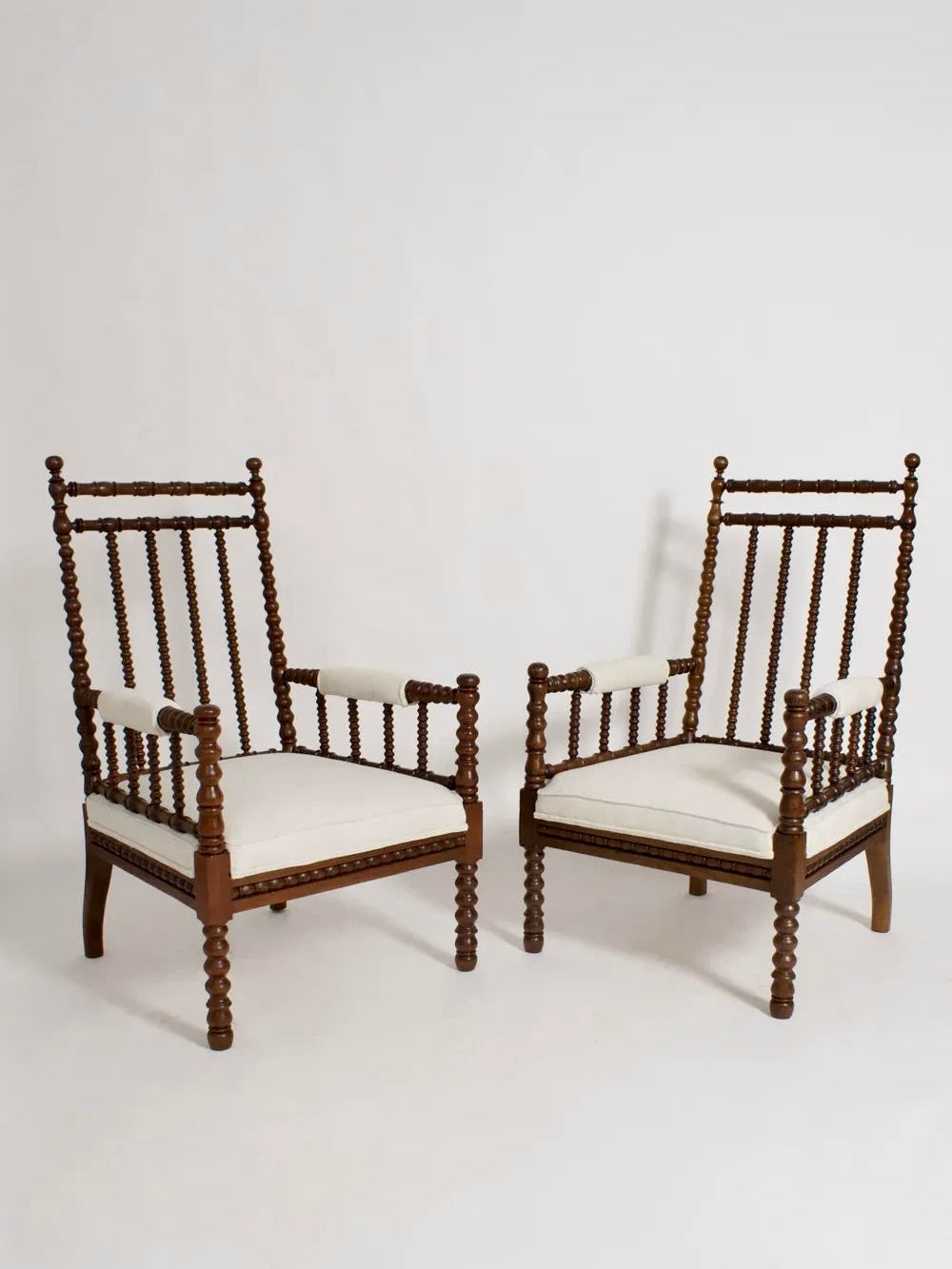 Barracuda Interiors offers a pair of 19th-century wood armchairs from England, featuring a spindle design with white cushions set against a plain background. These chairs showcase intricate detailing on the arms and legs, complemented by high backs that provide a traditional and elegant appearance.