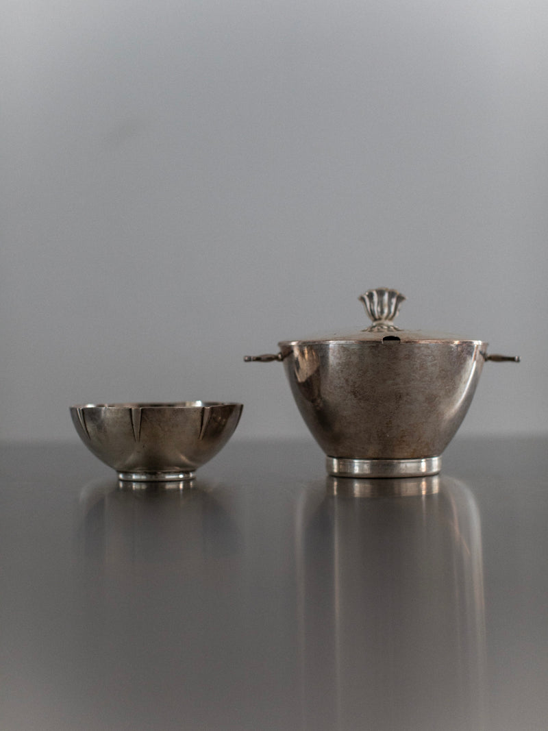 The Art Deco Bowls from Out For Lunch, featuring a shiny silver finish and a sugar bowl with a lid and handles, are displayed on a reflective surface. These bowls exude vintage glamour against the neutral gray background, adding a touch of timeless elegance to any setting.