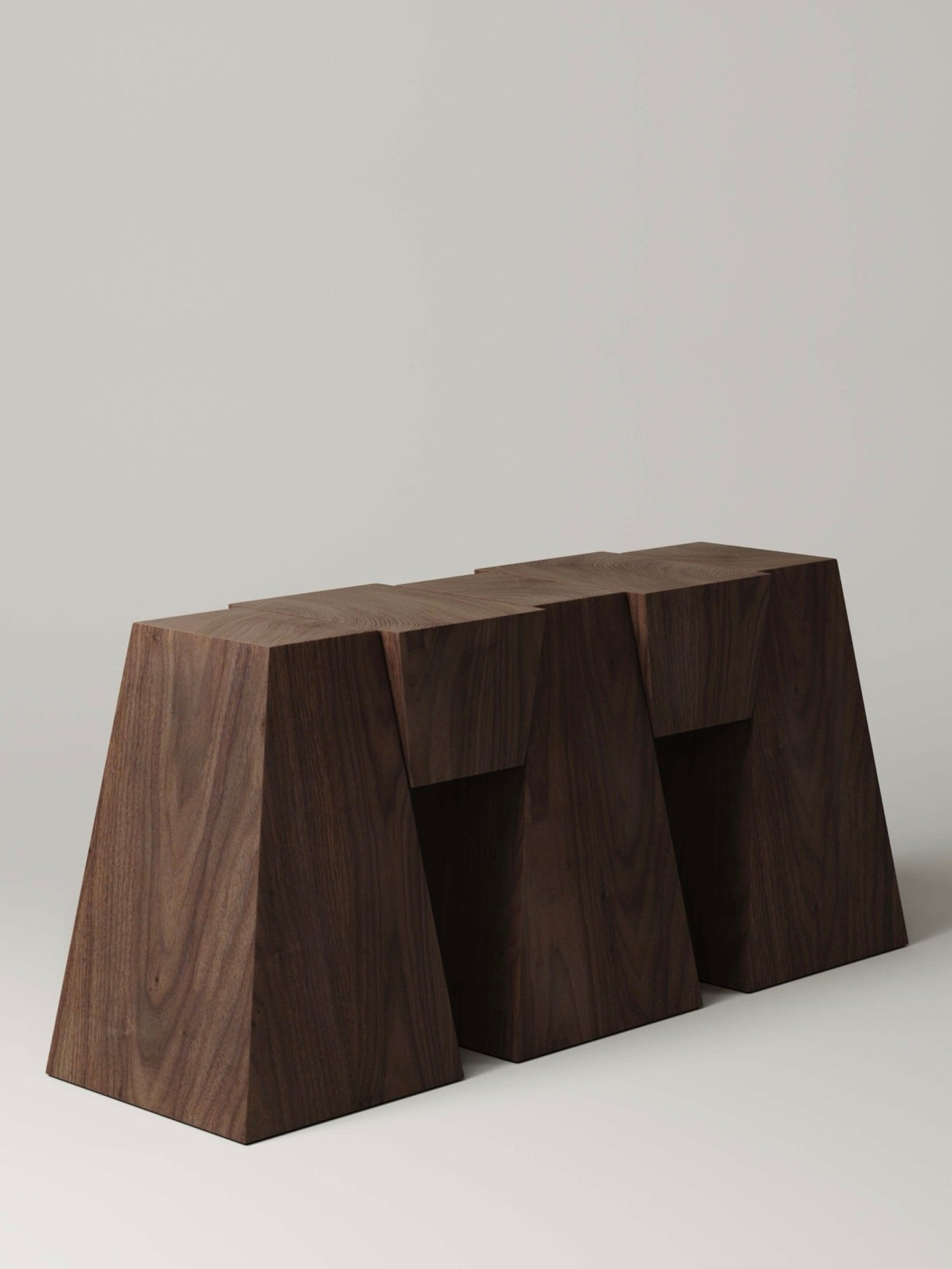 The M_006 Walnut Console by Monolith is a minimalist wooden bench featuring three connected trapezoidal shapes. The rich, dark finish complements its geometric, keystone-like design against a light background, enhancing its architectural elegance.