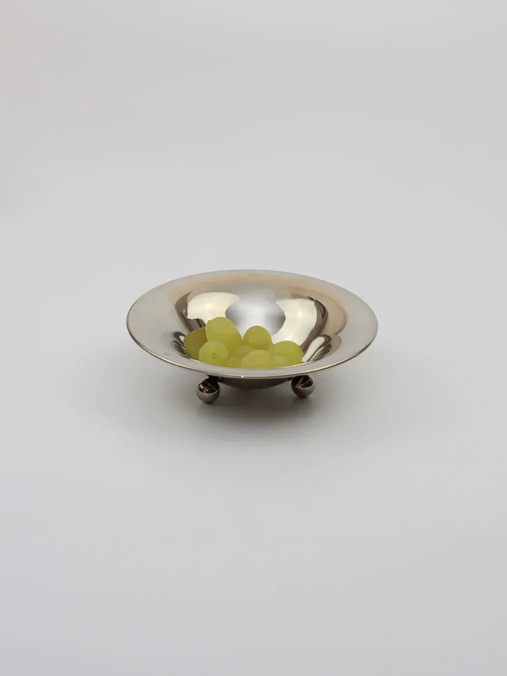 The Alpaca "Bowl on Three Legs" by Boga Avante Shop is small, silver, and reflective, holding a few green grapes against a gray background. Its design echoes the elegance of late 20th-century oyster serving.