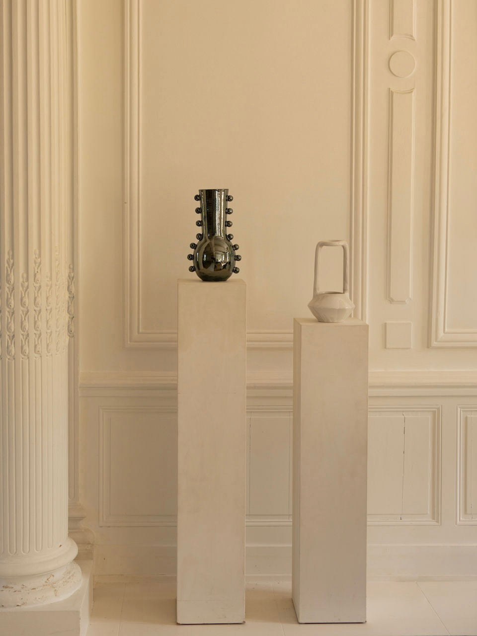 Two white pedestals stand in a minimalistic room with ornate wall details. One pedestal holds the Vase Solstice II from Landy Rakoto Ceramics, featuring a reflective silver metallic effect finish; the other, a sculptural ceramic white abstract jug. A decorative column is partially visible on the left.