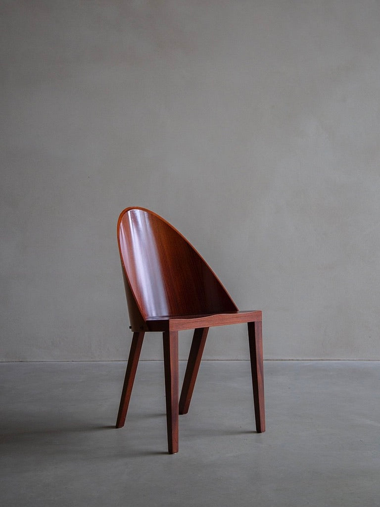 Against a plain gray wall, the Philippe Starck - Royalton 44 chair by Bicci de' Medici sits elegantly with its minimalist style, curved backrest, four straight legs, and rich brown wood finish reminiscent of the Royalton Hotel design.