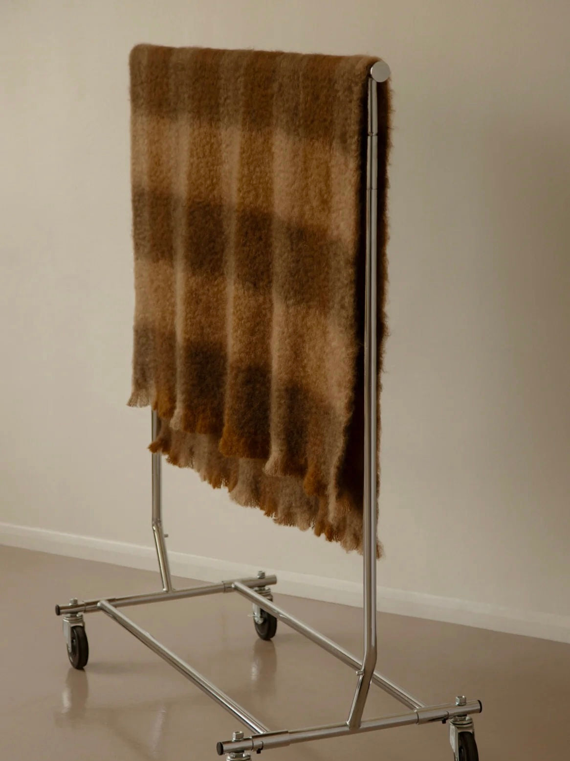 An "Arbor Sable" Throw from The Wren, a luxurious brown plaid blanket made with eco-friendly mohair, is neatly draped over a metal clothing rack with wheels, framed by a plain beige wall and floor.