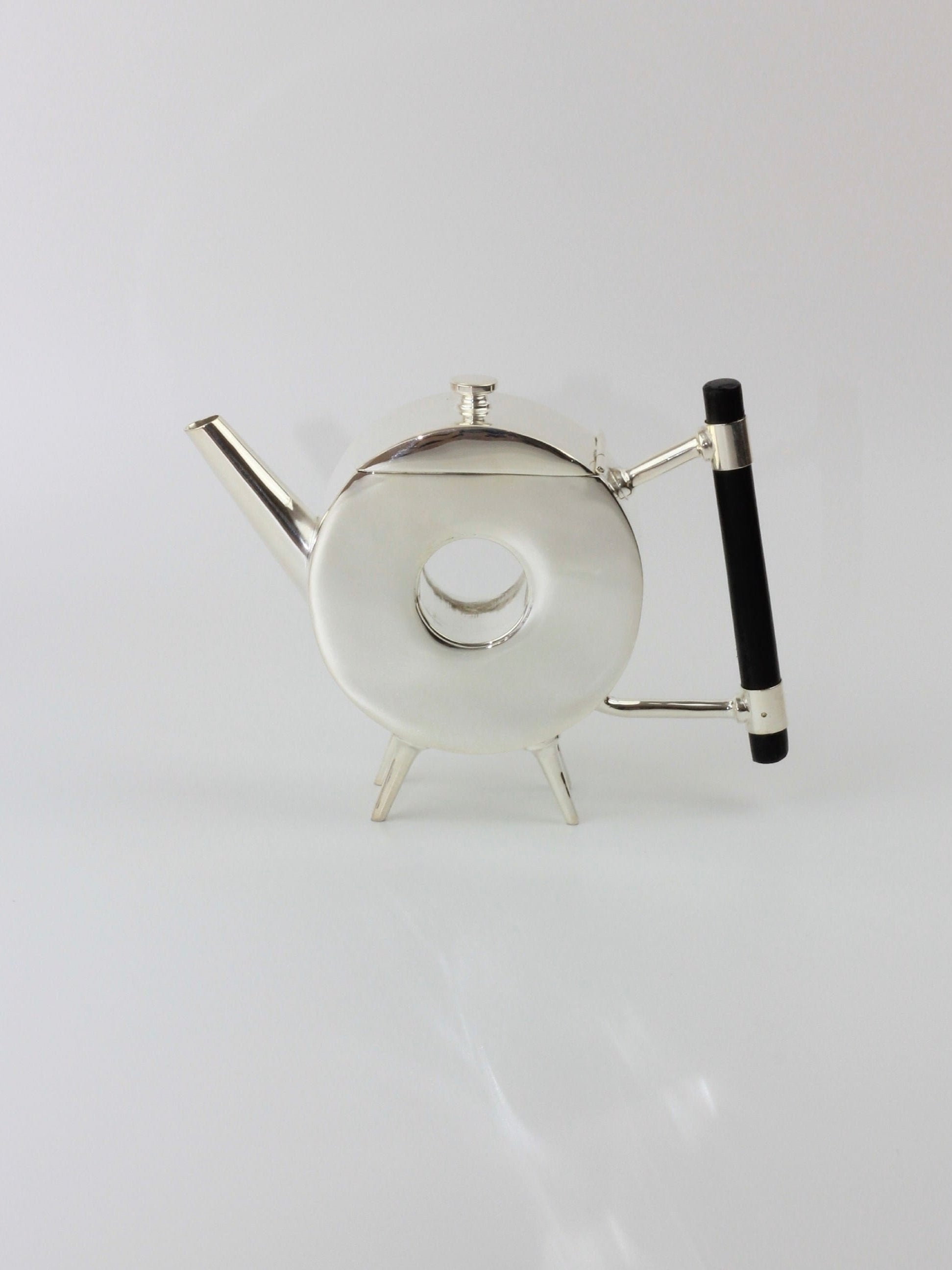 The Christopher Dresser's Tea Pot from Boga Avante Shop is a sleek, modern teapot featuring a unique round, hollow center design. Its silver-plated finish pays homage to Dr. Christopher Dresser's aesthetic, and it includes an ebonized wood handle on the right. The left-side spout and three small legs further enhance its contemporary and artistic appearance.