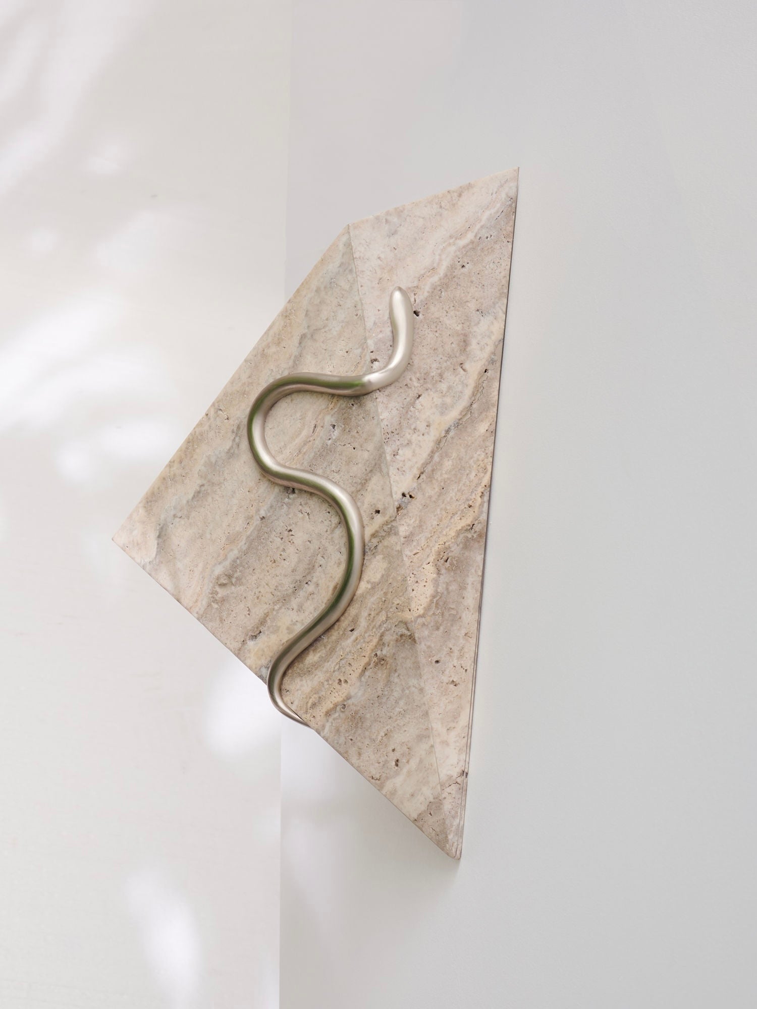 The Mariza Galani's Therapeutic Wall Light 3 is a triangular, marble-like wall sculpture featuring a smooth, winding metallic element that extends diagonally, resembling a therapeutic sconce. This exquisite piece casts soft shadows on the light-colored background, enhancing the depth and dimension of its minimalist design.