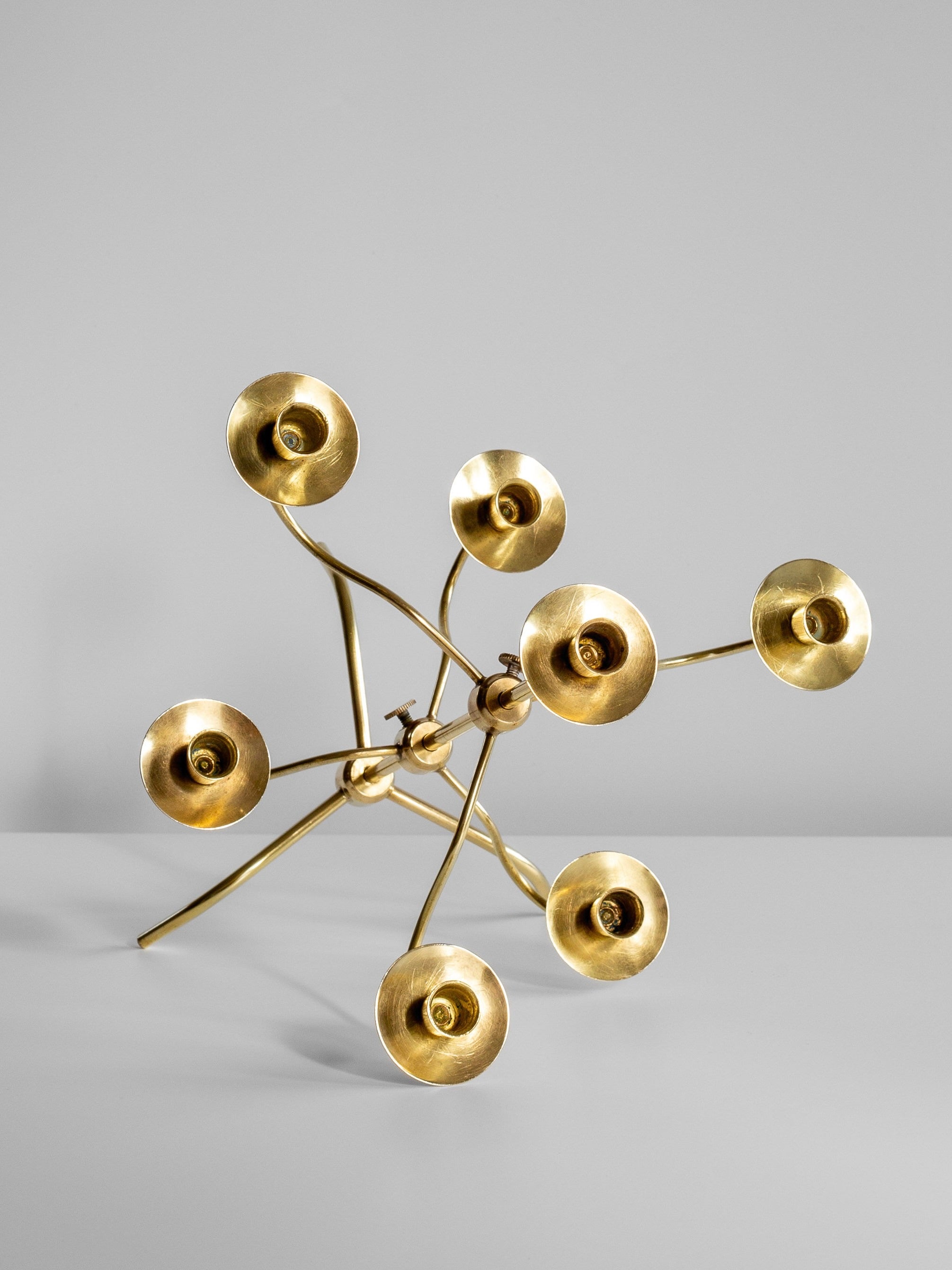 The Medium Mid-Century Brass Candelabra by Spigel is a modern, abstract piece reminiscent of Svenskt Tenn designs, featuring seven circular, flower-like structures connected by slender, curved rods. The candelabra showcases an organic, free-form design and stands on a plain surface against a neutral background.