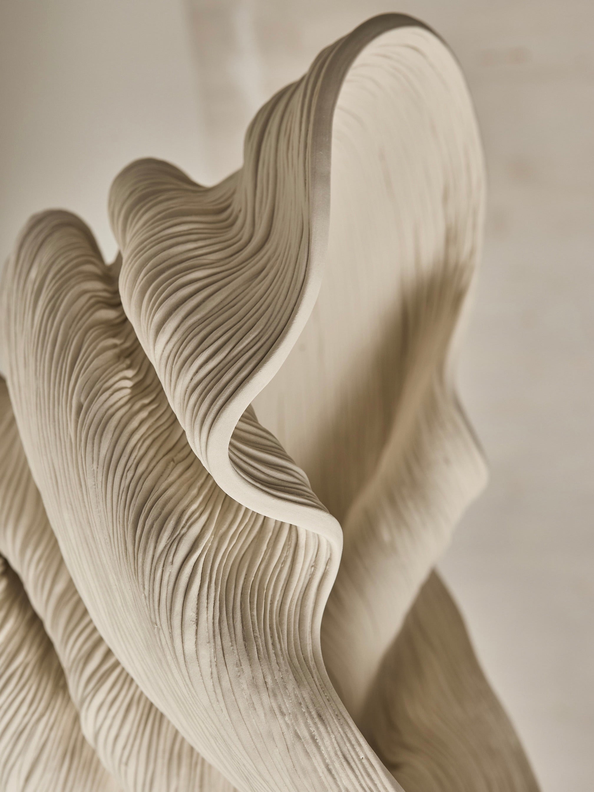 Close-up of the Medium Flutter by The Soucy Shop, a unique, handmade abstract sculpture crafted from porcelain coils. This off-white piece features wave-like forms and ribbed textures with smooth curves and intricate details resembling a natural structure against a softly blurred background.