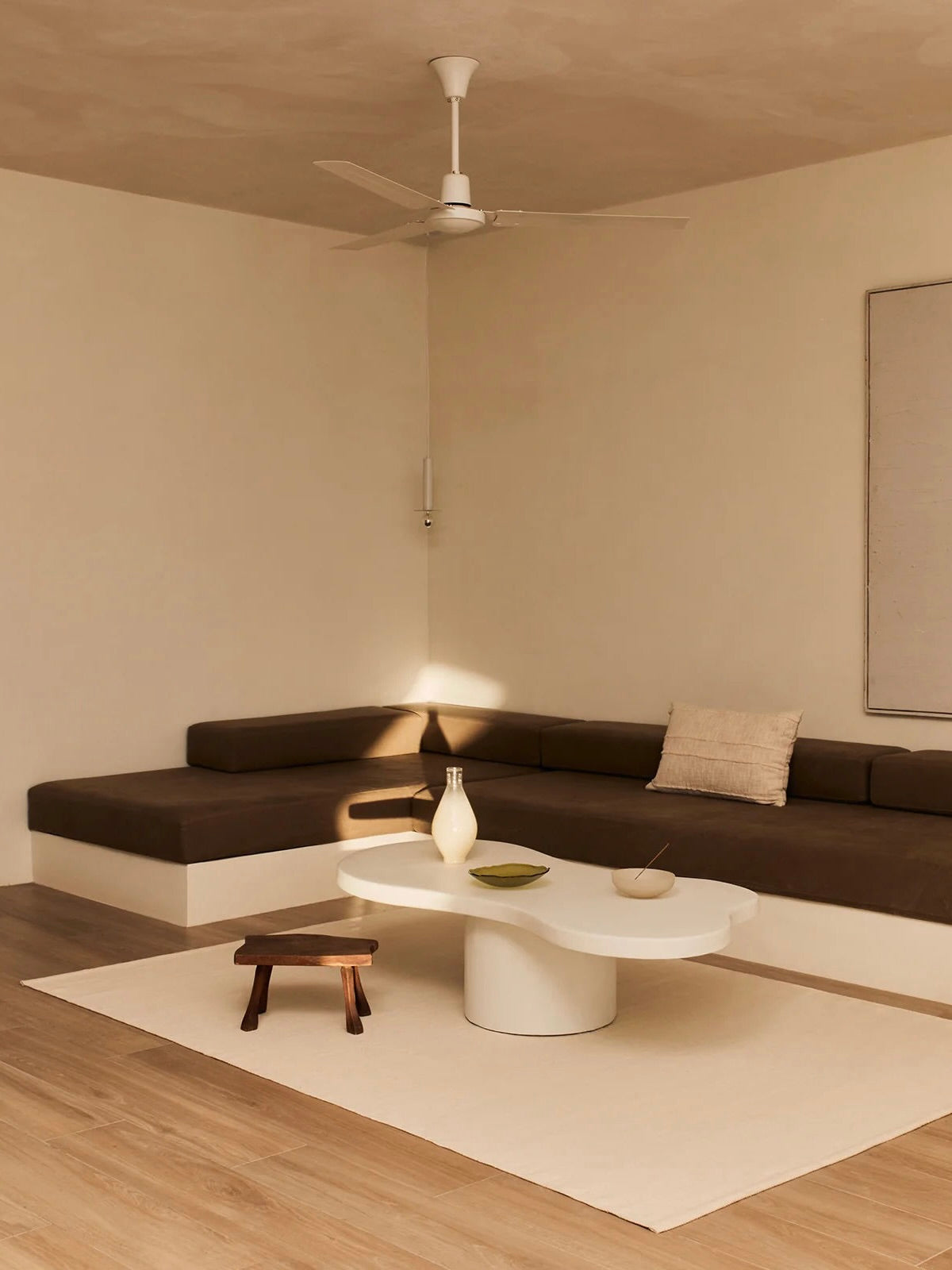 A minimalist living room showcases a brown L-shaped sofa paired with the "Pesto" Coffee Table by MarlotBaus, known for its artisan-crafted, oval shape. A small wooden stool complements the white ceiling fan, beige rug, vase, and bowl on the table. A rectangular wall art piece hangs above the sofa.