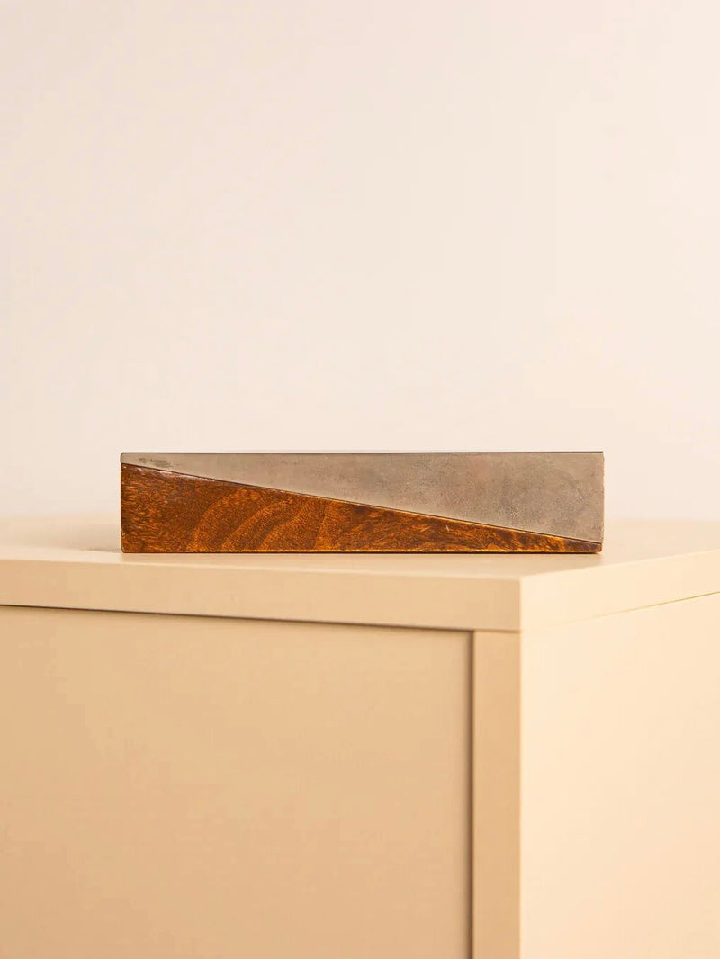 The Square Vide-poches 60's by Treaptyque, a minimalist decorative block made of wood and metal featuring a diagonal design with a stunning patina, sits elegantly atop a beige cabinet against a plain wall.