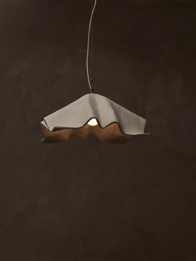 The 'La Fleur' Pendant Light by Studio Tristan Kallas features a wavy, scalloped-edged shade that casts warm, dimmable light against a dark, textured background, creating an elegant and modern minimalist atmosphere.