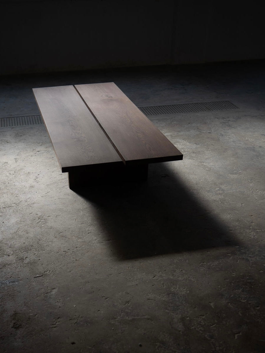 The Tanizaki Coffee Table by ASERIES OF OBSTACLES, SL, is a minimalist rectangular wooden table inspired by Japanese living rooms. Sitting close to the ground, its sleek structure features two adjacent wooden planks casting a long shadow on the dimly lit concrete floor.