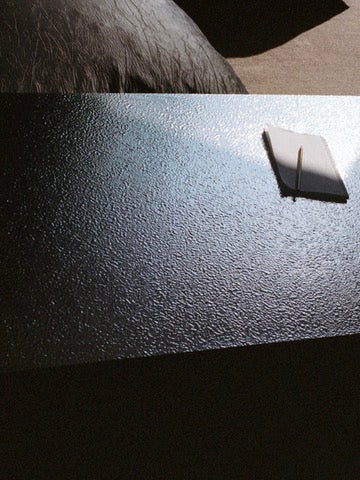 A silver Mara Sister coffee table by TEGET is partially lit by sunlight, highlighting a small, closed white notebook with a pen on top. The background remains dimly lit, emphasizing the contrast of light and shadow.