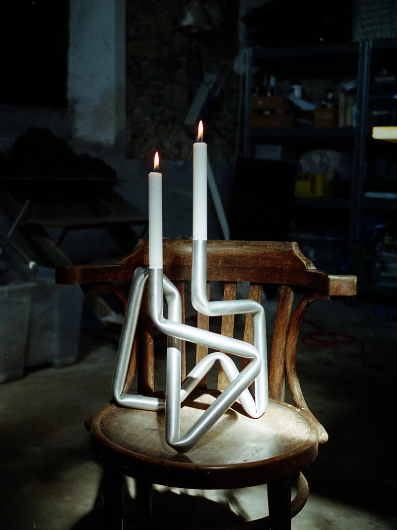 A pair of white candles lit atop an AOT STUDIO hand-made Bucati Candle Holder (brushed aluminium) sits on a weathered wooden chair in a dimly lit room filled with various tools and shelves in the background.
