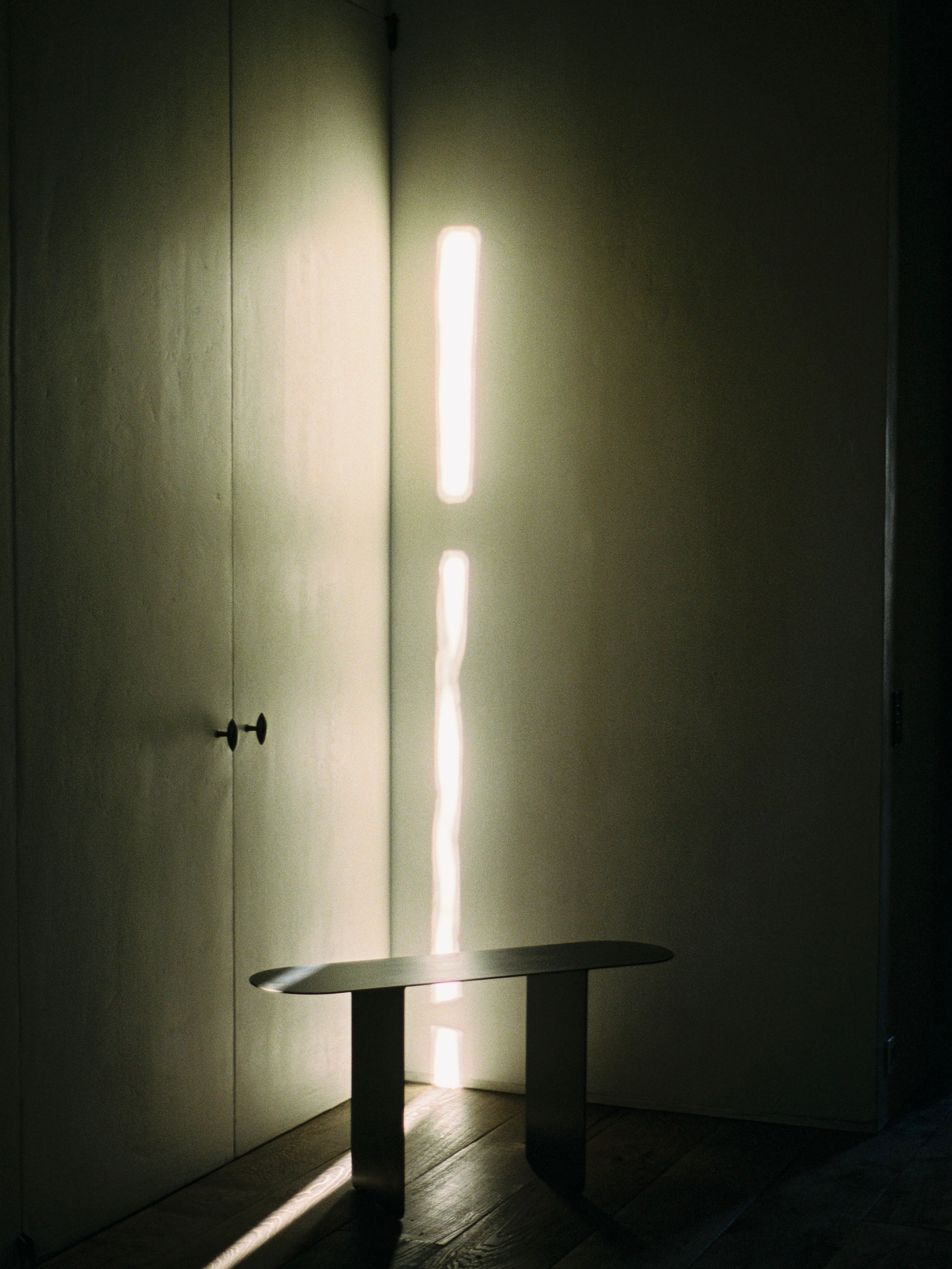 Sunlight streams through a narrow vertical window onto a dark wall, creating elongated light patterns. In the foreground, "The Altar" by COSEINCORSO, a minimalist bench akin to prayer kneelers, sits illuminated by the sunlight and casting shadows on the wooden floor.