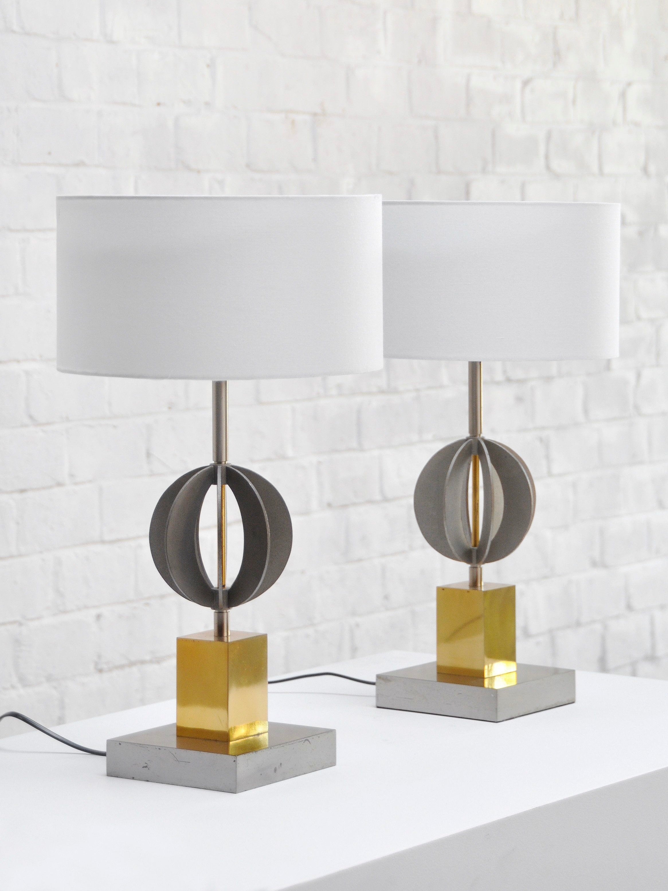 The Pair of Italian Geometric Table Lamps 70's by Introverso features chrome and brass geometric designs with white cylindrical shades on yellow rectangular bases, elegantly set against a white brick wall.