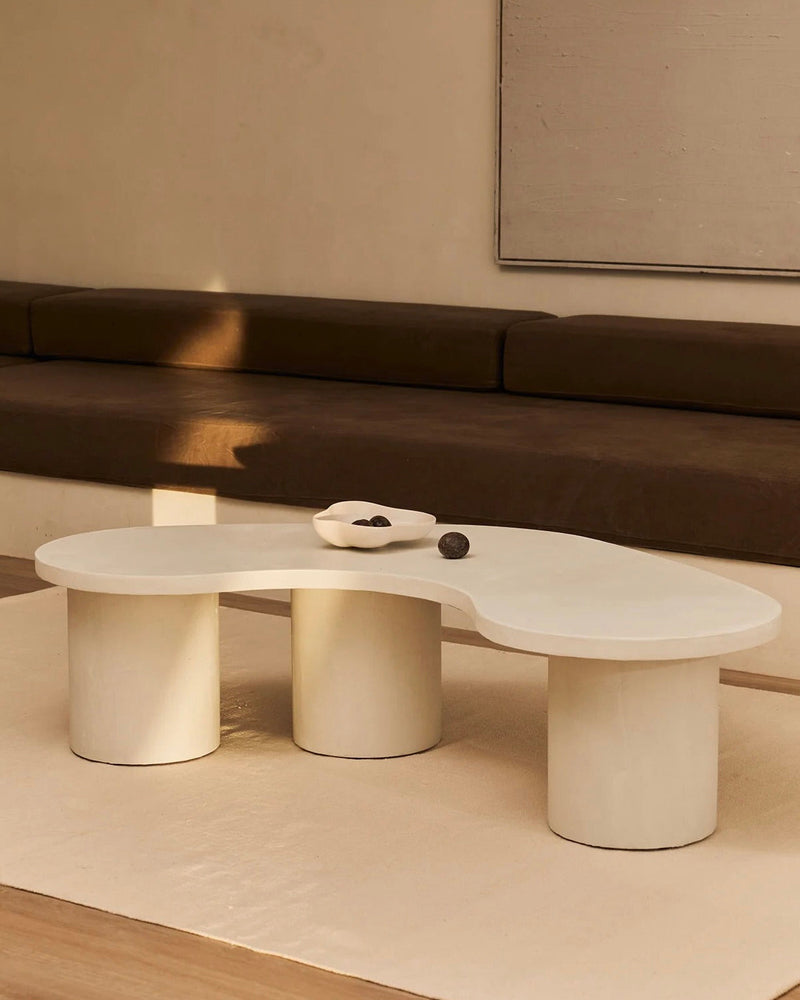 In a minimalist living room, MarlotBaus's "Parmigiano" Coffee Table boasts an abstract wavy shape with cylindrical legs. A small white dish with decorative stones sits on the table, while a beige couch with brown cushions completes the sophisticated look against a neutral microcement wall.