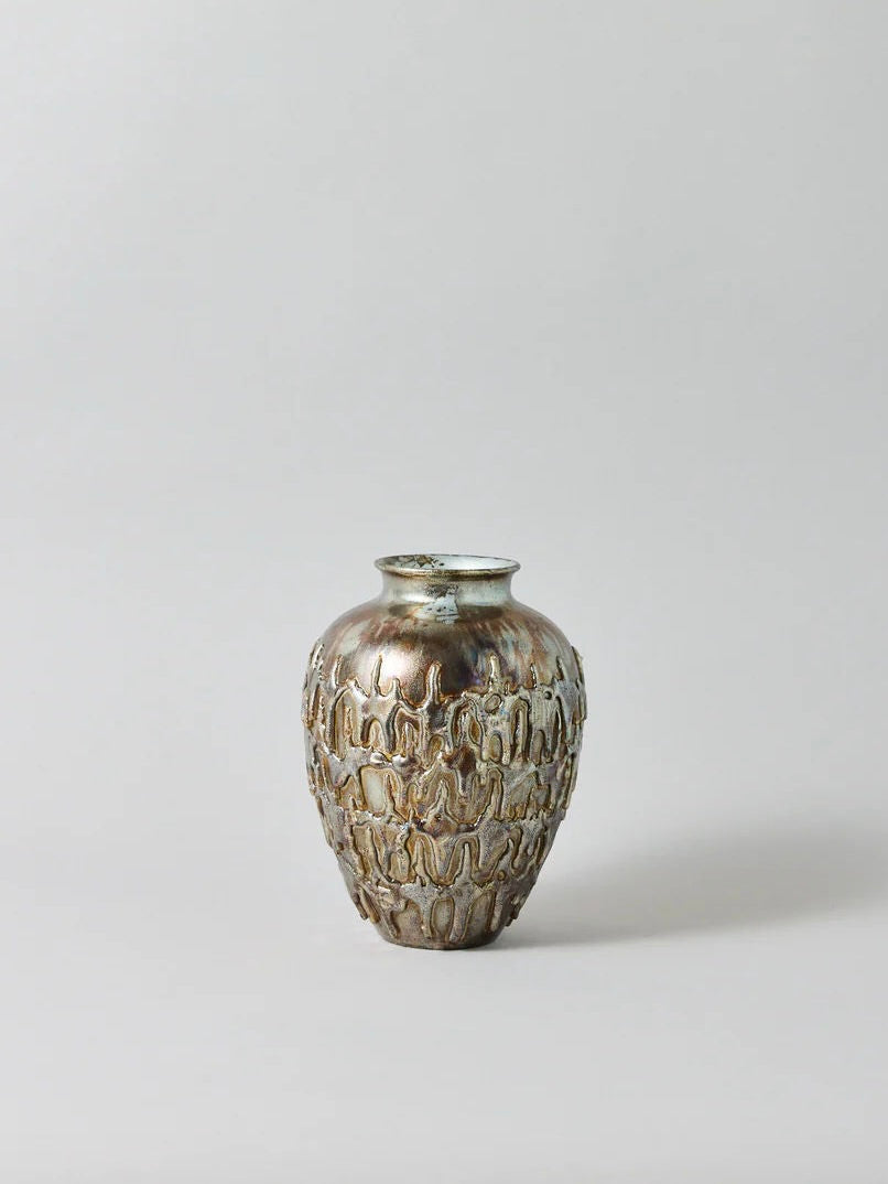 The "Almost a Mirror" Vase by John Nordenstein is a distinctive, handmade decorative ceramic piece. It boasts a narrow neck and wider body, adorned with an intricate, raised pattern reminiscent of dripping wax. Finished with a metallic silvery-bronze glaze against a plain, light gray background, it truly emphasizes its unique design.