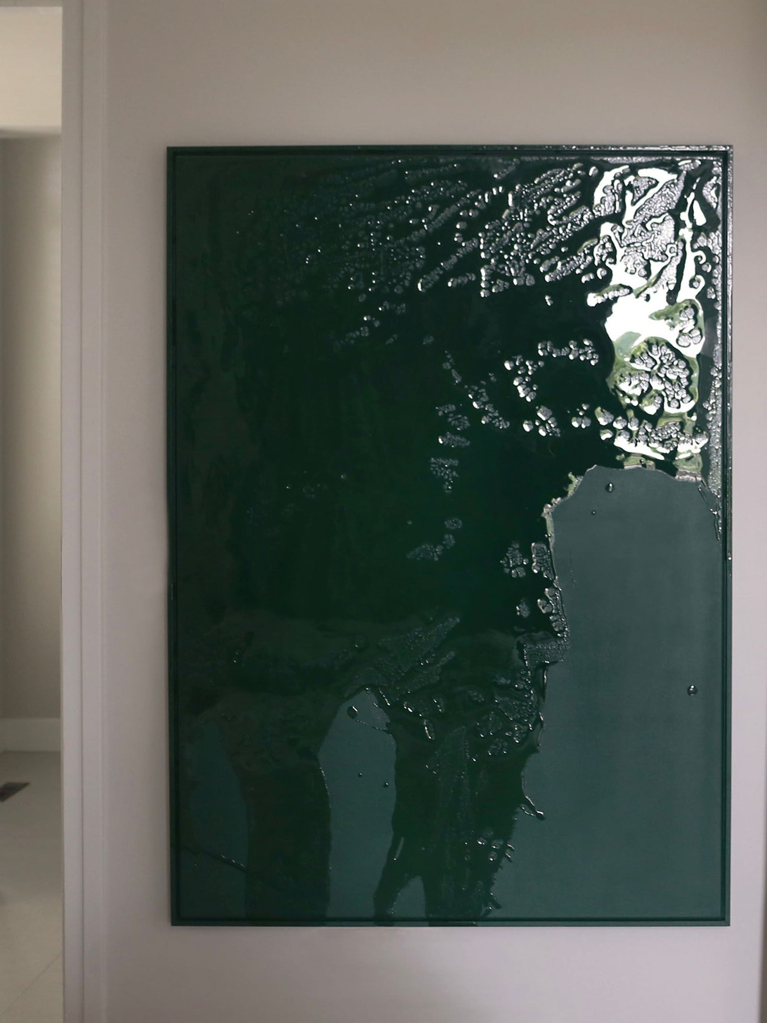 The "Green Flow" painting by STUDIOPOLS decorates the wall, featuring a dark green textured abstract design crafted with acrylic paint. Its glossy finish and uneven patterns produce a reflective sheen, giving depth to the artwork. This framed canvas is set in an understated black frame, amplifying its presence in a room with neutral tones.