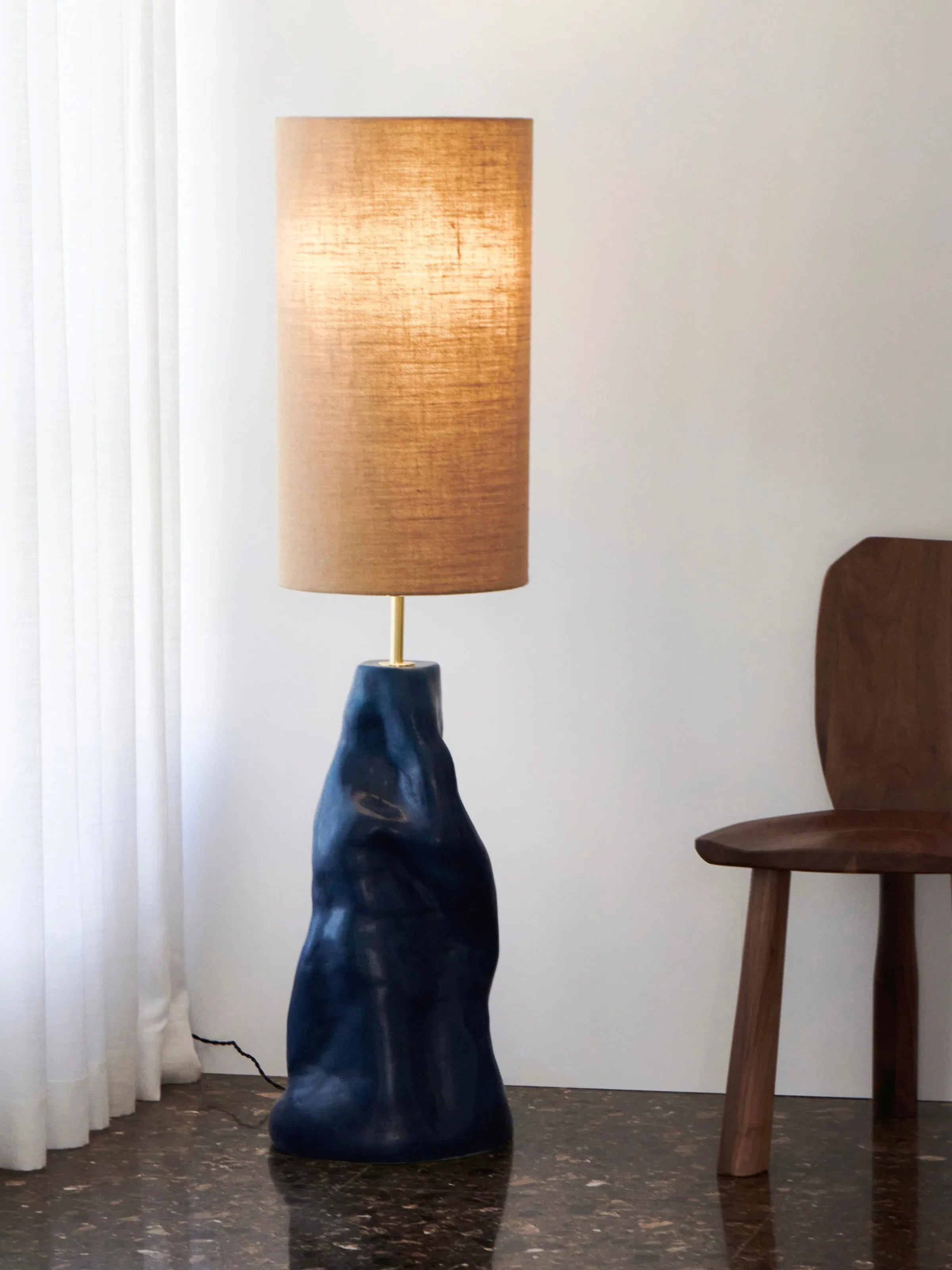 The "Menina" Floor Lamp in Blue by Project 213A is a modern fixture with an artisanal ceramic base, handcrafted in Northern Portugal. The lamp showcases a textured dark blue sculptural design, paired with a tall cylindrical beige fabric shade. It stands elegantly on a glossy dark marble floor next to a wooden chair, casting a warm and cozy glow that harmonizes perfectly with white curtains and the plain wall in the background.