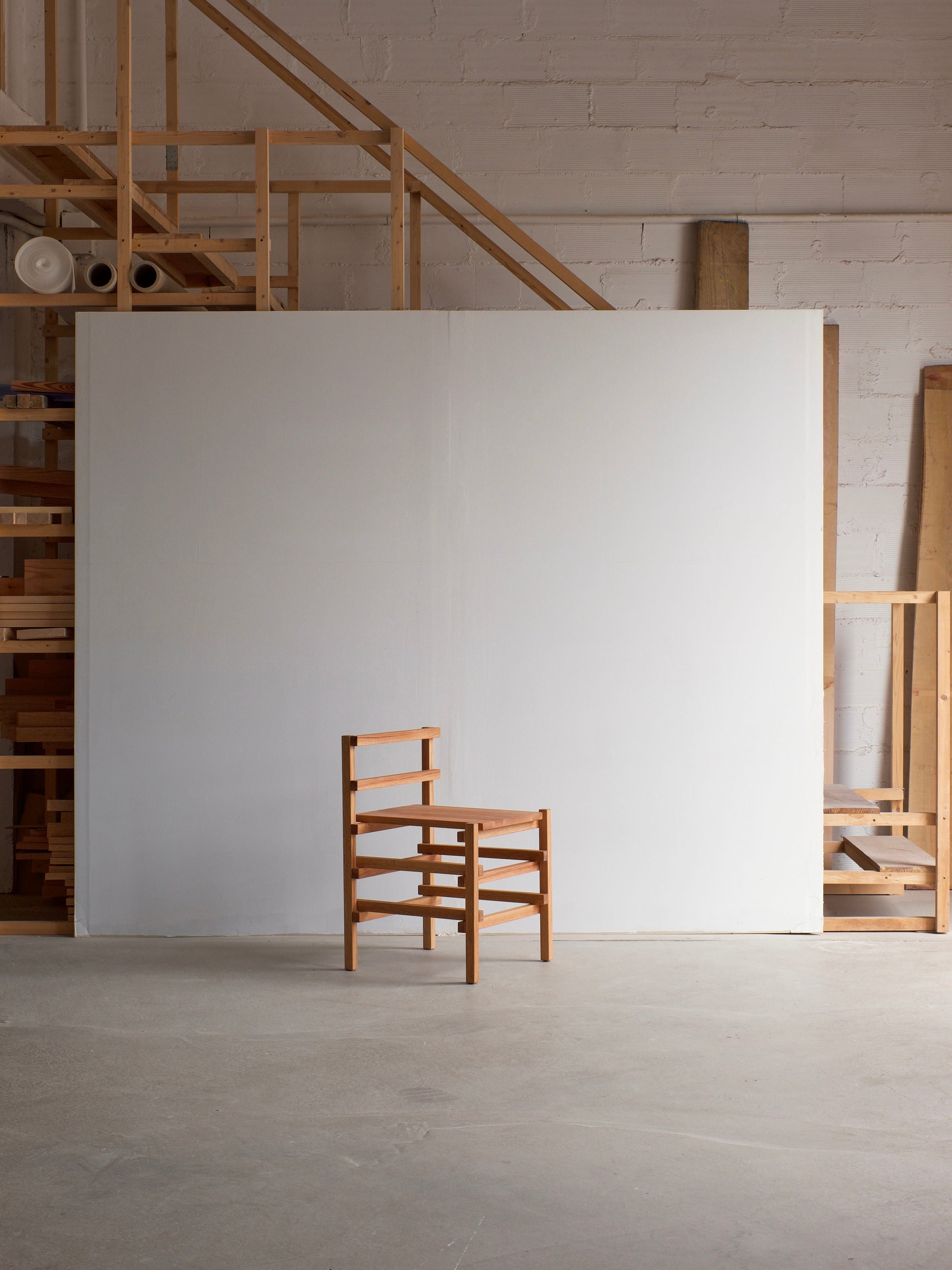 A minimalist Small Palma Chair by malva, featuring a ladder-like backrest, is positioned in front of a large white panel. The setting includes a wooden framework staircase that ascends to a loft, accompanied by various wooden materials and tools nearby, crafting a versatile statement piece within the workshop's ambiance.